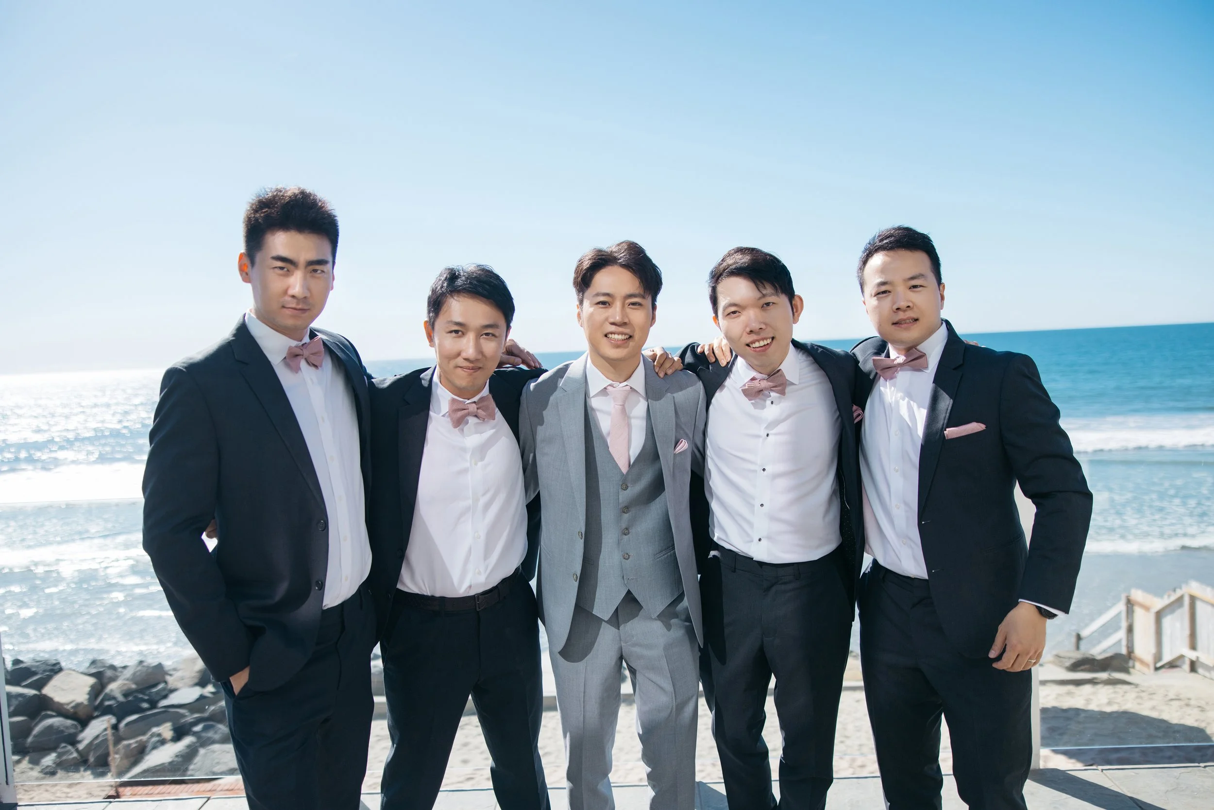 Five young men in suits with pink bow ties standing together on a beach, smiling, with the ocean and blue sky in the background.