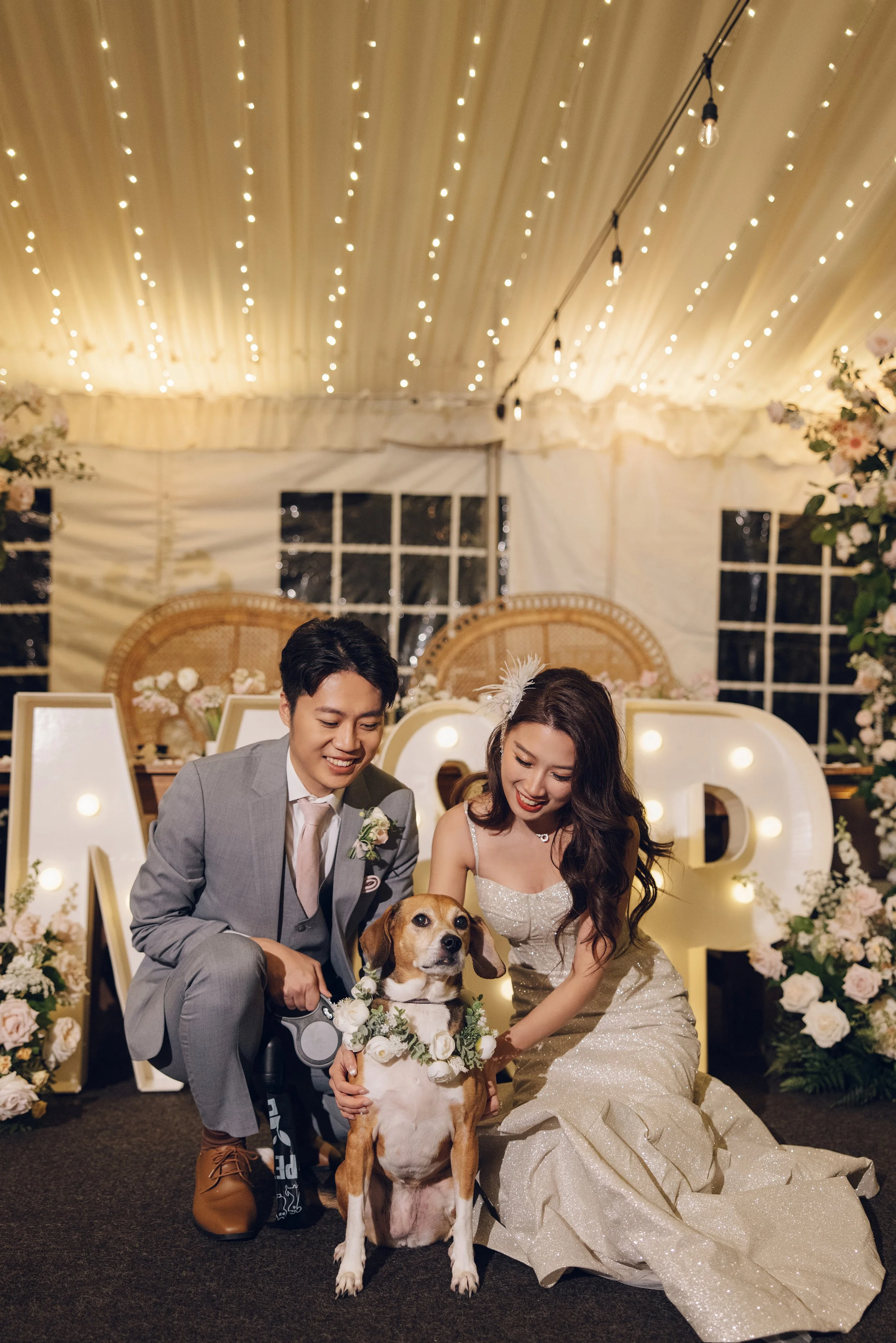 A newlywed couple with their dog at a wedding reception in a decorated tent, with string lights and illuminated marquee letters in the background.