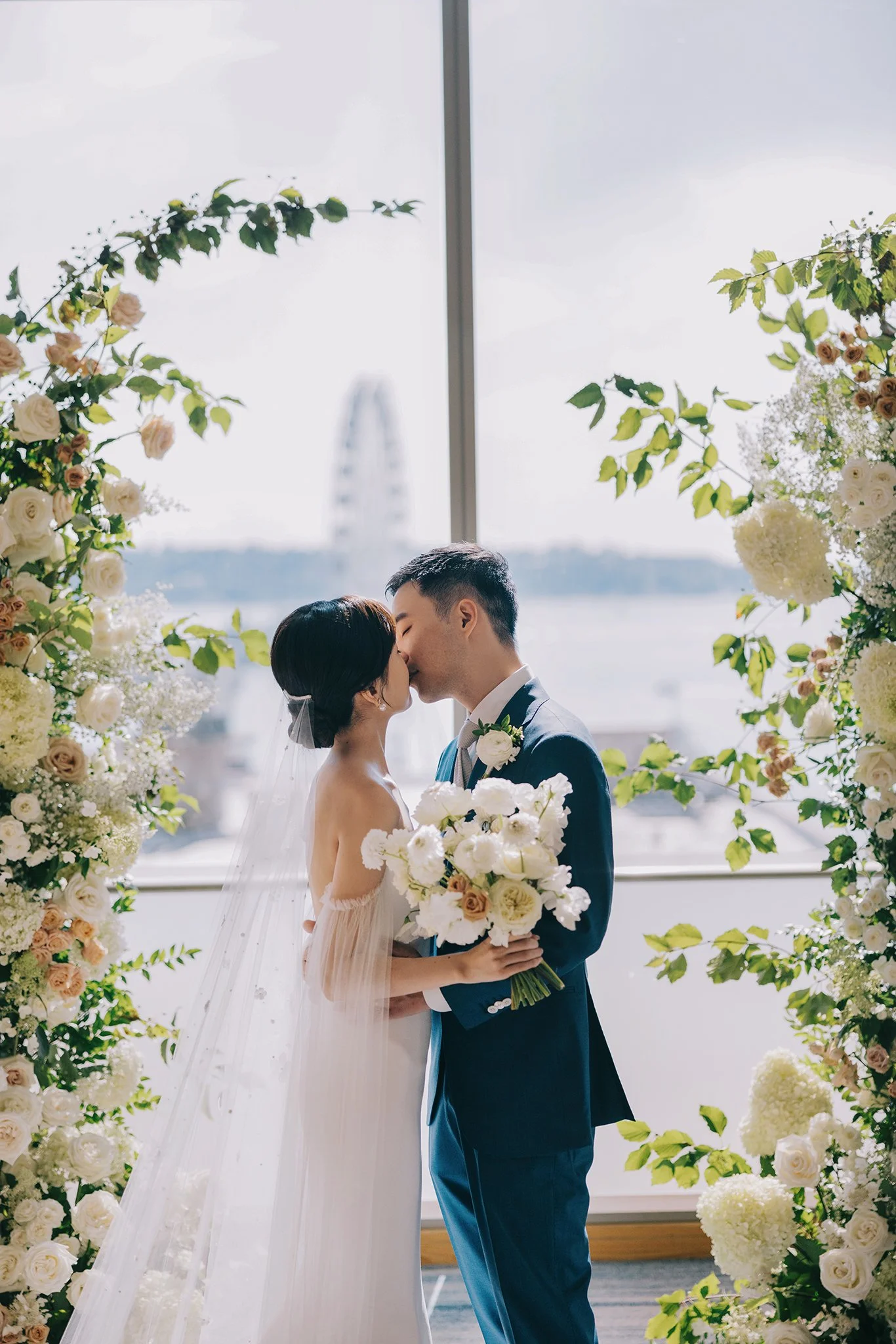 A bride and groom are kissing at their wedding, surrounded by floral arrangements, with a large window behind them showing a Ferris wheel and water.