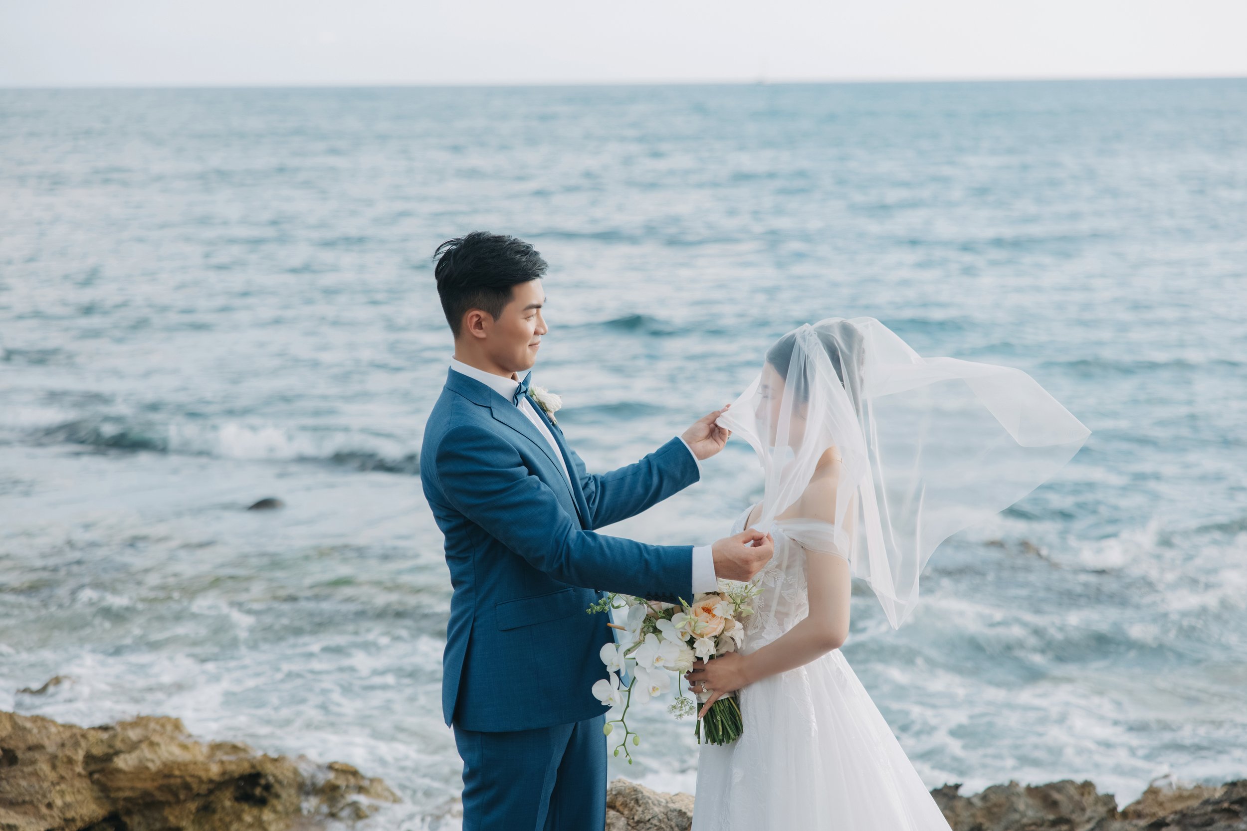 A bride and groom standing on rocky shore at the ocean, with the groom lifting the bride's veil.