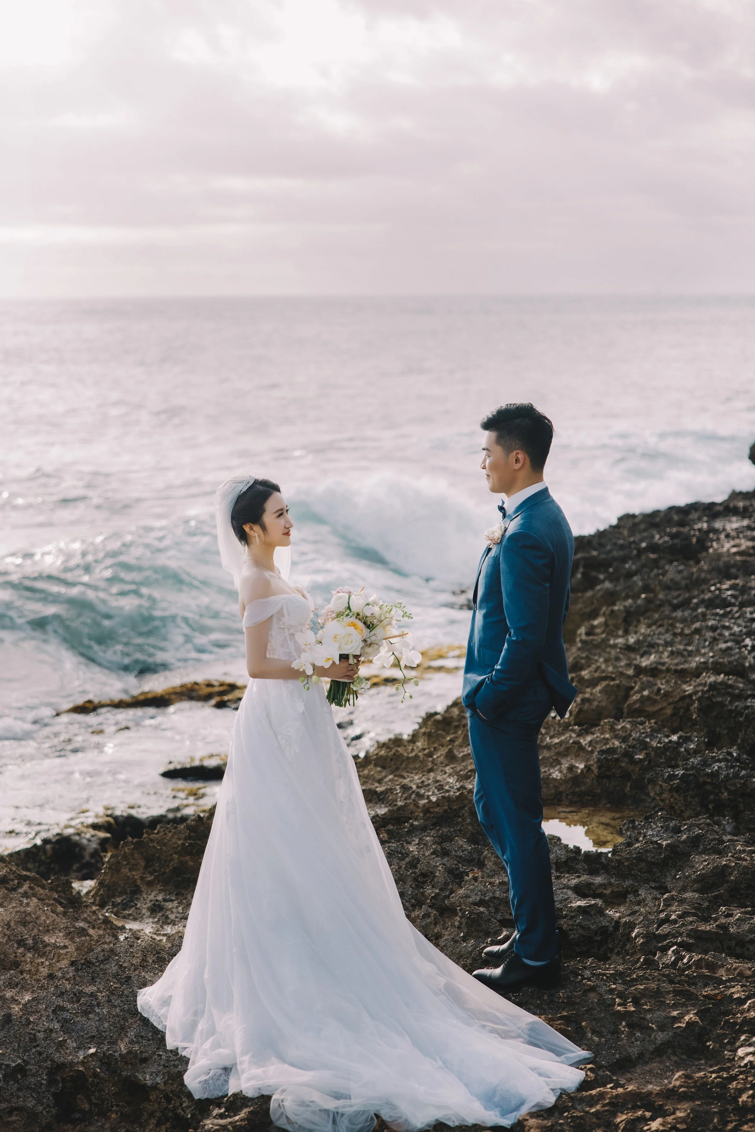 A bride in a white wedding dress holding a bouquet standing on rocks by the ocean, facing a groom in a blue suit, during sunset.