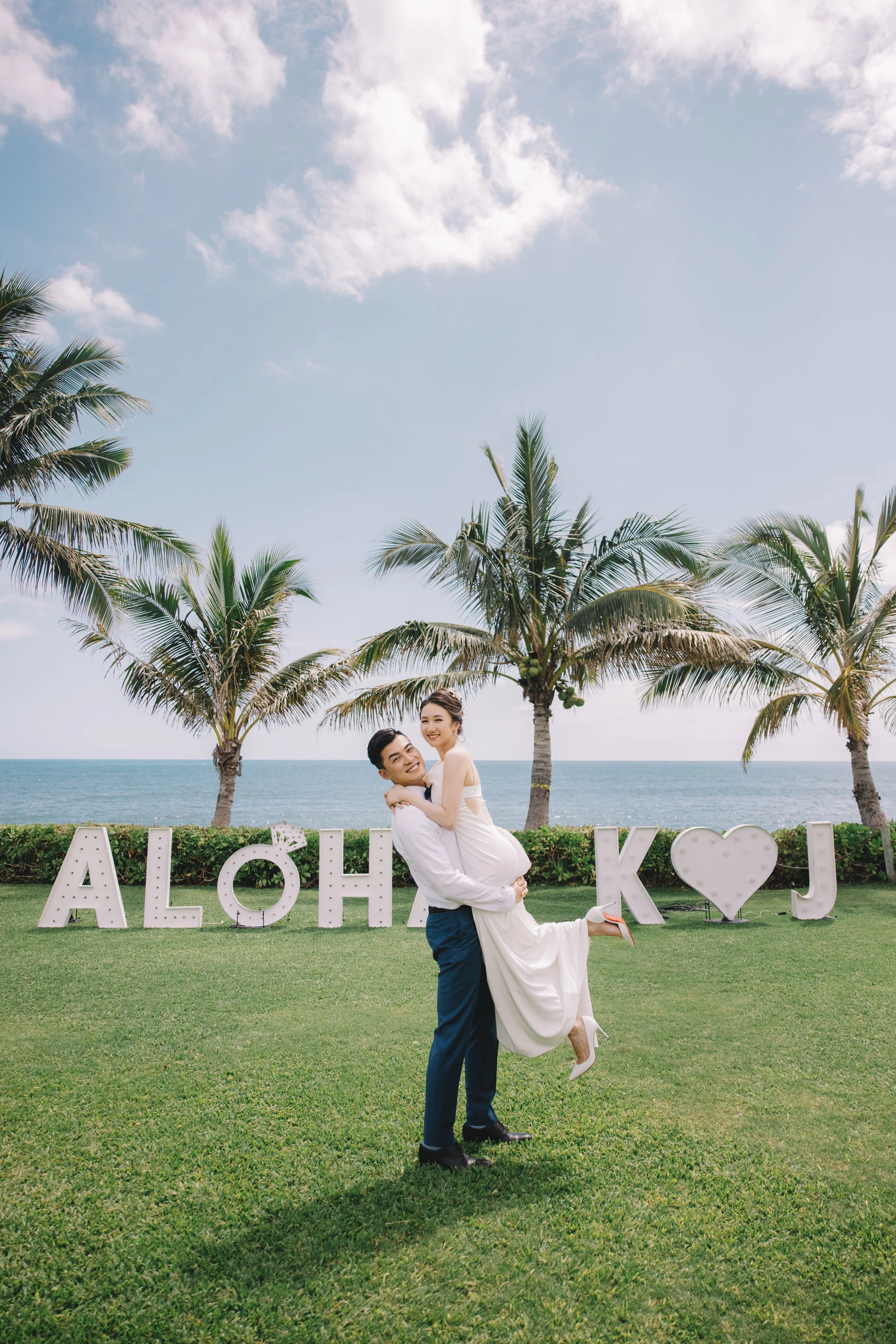 A couple dressed in wedding attire, with the groom lifting the bride, standing on a grassy area with palm trees and the ocean in the background. Large decorative letters spell out 'Aloha K J' behind them.