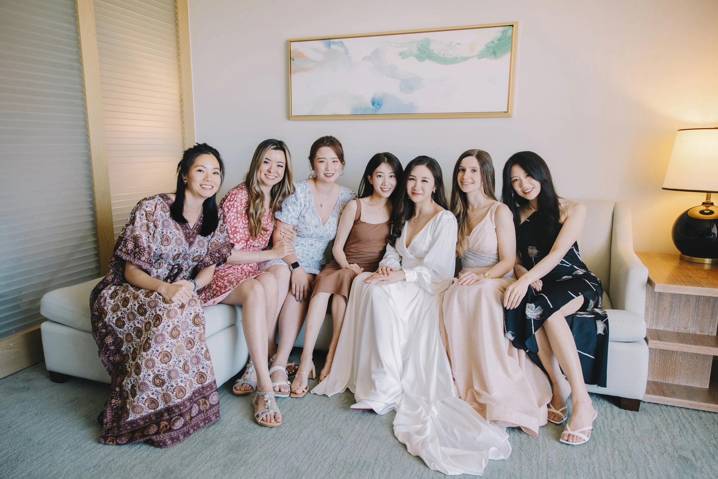 A group of eight women sitting on a beige sofa in a living room, dressed in stylish dresses with a large abstract painting on the wall behind them and a table with a lamp on the right.