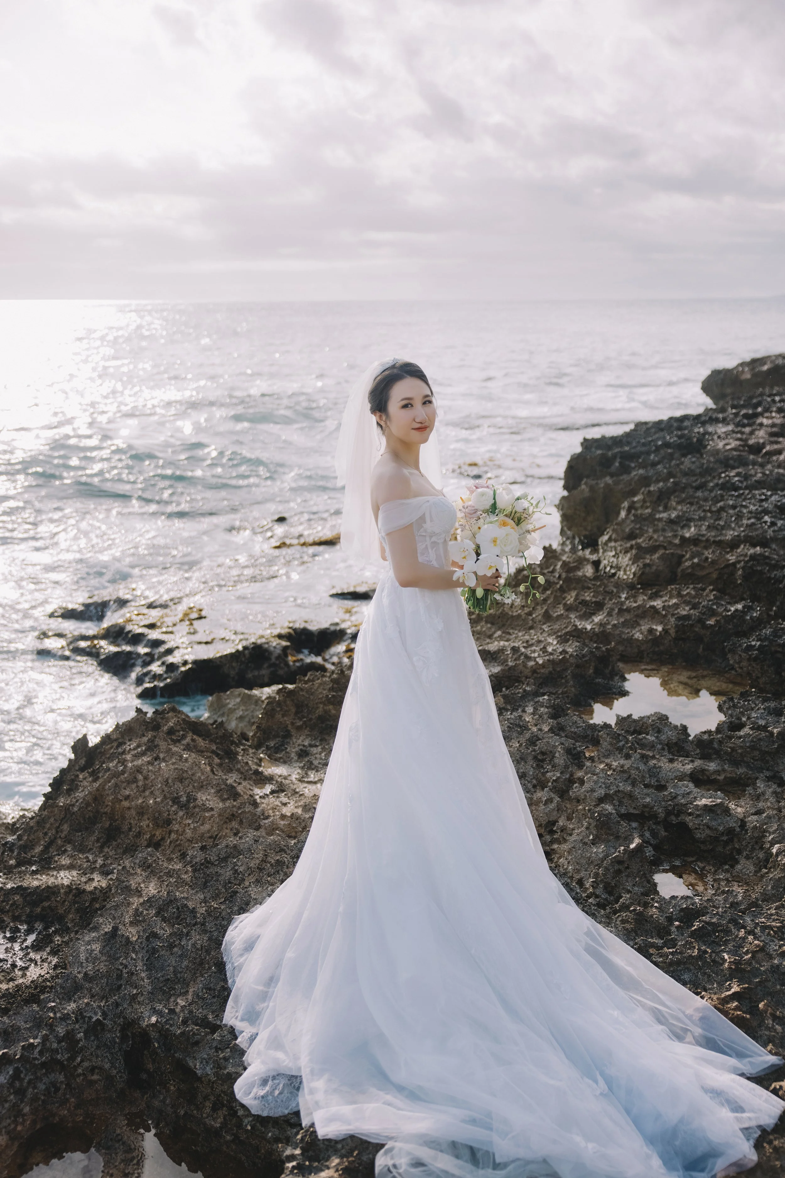 A bride in a white wedding dress and veil holding a bouquet of flowers on a rocky shoreline near the ocean during daytime.