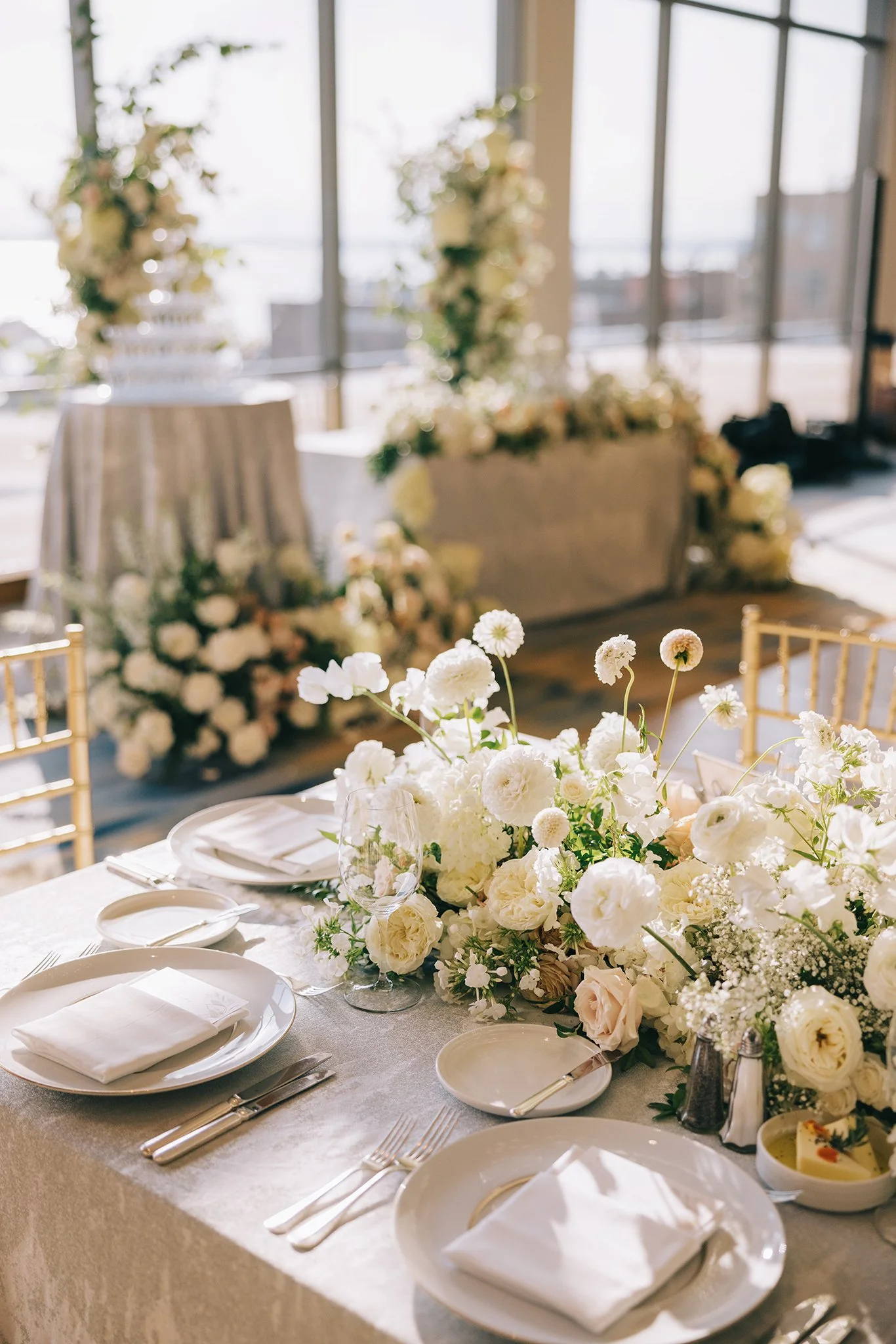 Elegant wedding reception table setup with white floral centerpiece, white plates, napkins, silverware, wine glasses, and gold chairs, with large windows and floral decorations in the background.