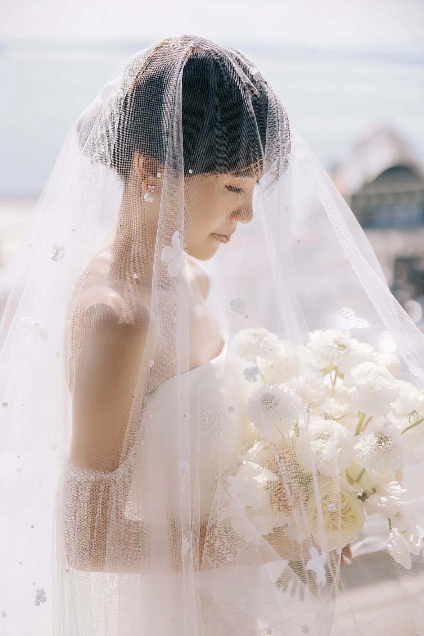 A bride in a wedding dress and veil, holding a bouquet of white flowers, smiling softly with her eyes closed, outdoors on a bright day.