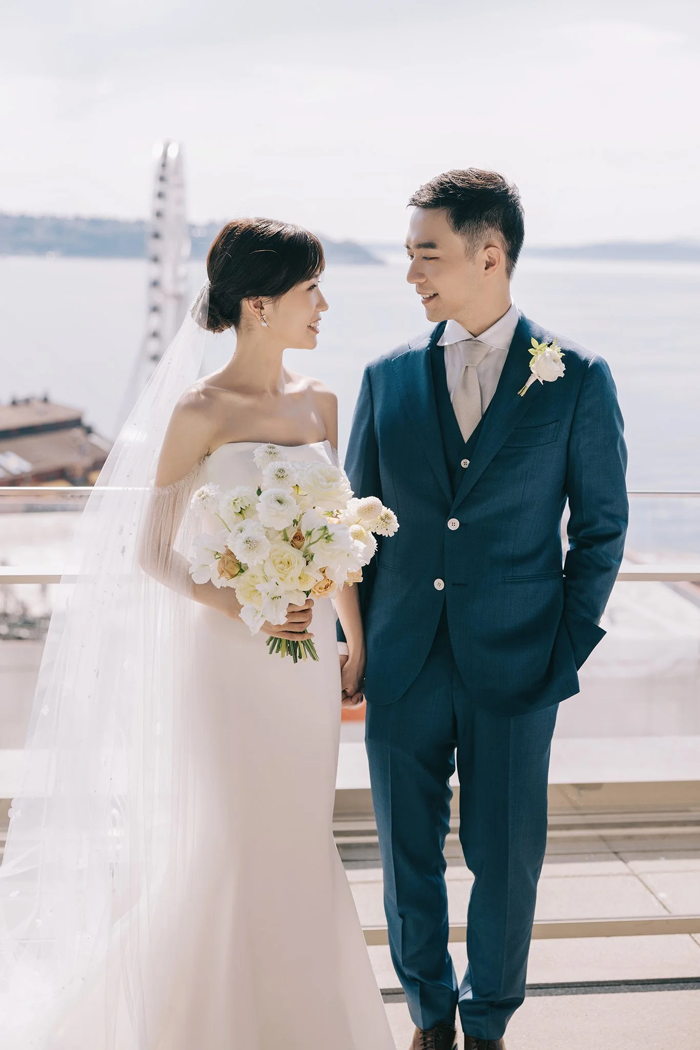 A bride and groom standing on a balcony overlooking a body of water, sharing a moment during their wedding. The bride wears a white off-shoulder wedding dress and holds a bouquet of white flowers. The groom wears a blue suit with a white shirt and li