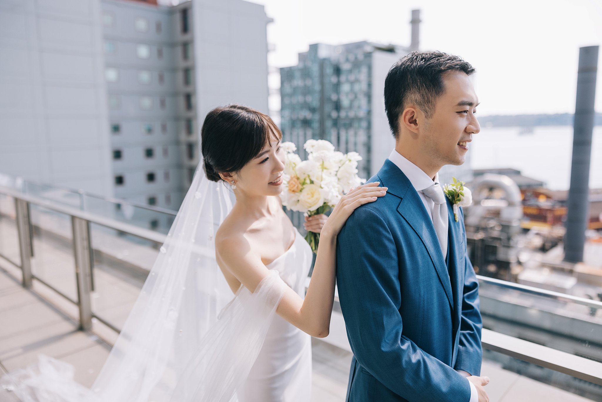 A bride and groom on a balcony in an urban setting. The bride is placing a hand on the groom's shoulder and holding a bouquet of white flowers, both smiling and looking forward.