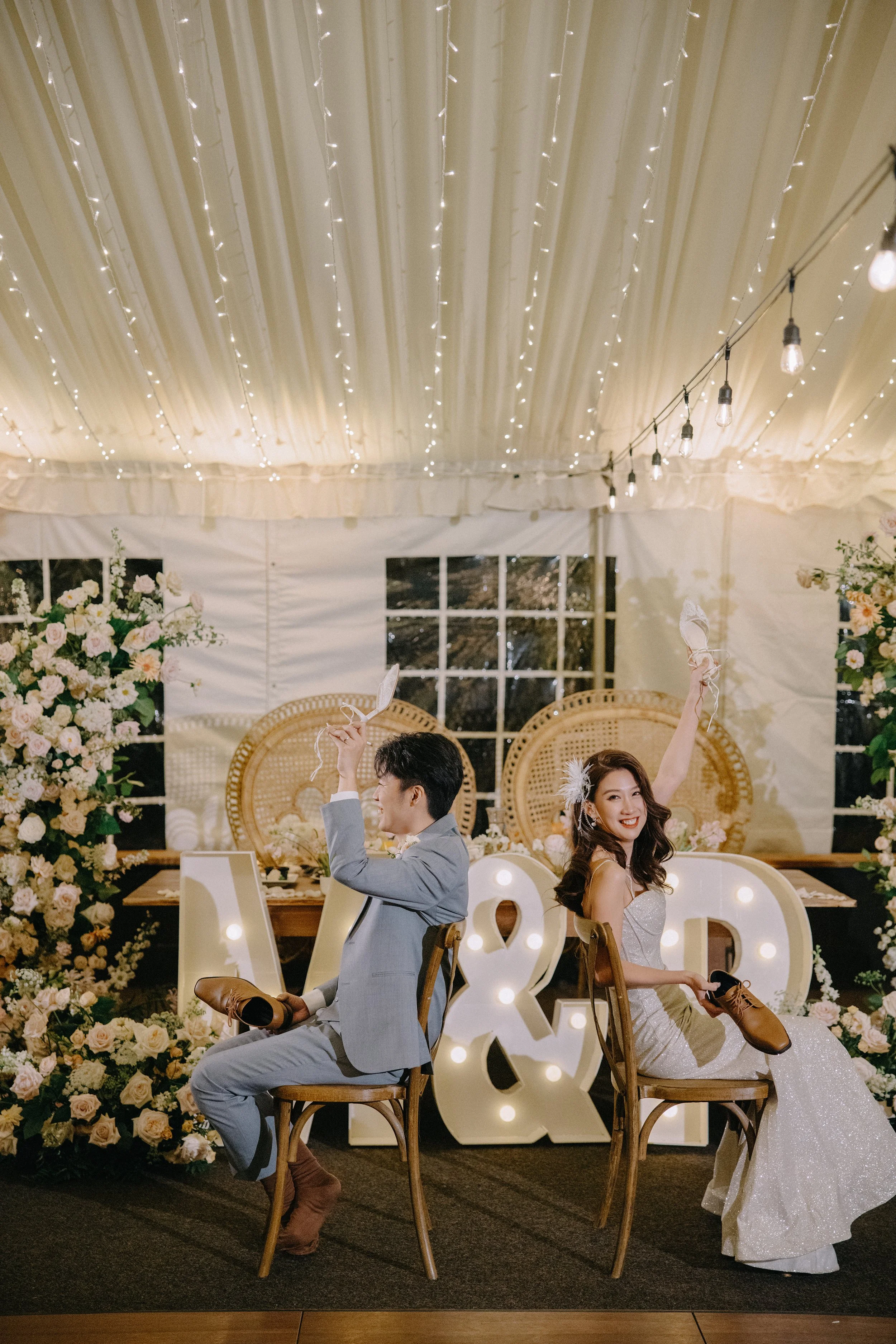 A bride and groom participating in a shoe game at their wedding reception, sitting back-to-back in a decorated venue with flowers, fairy lights, and illuminated letters that spell "A & R."