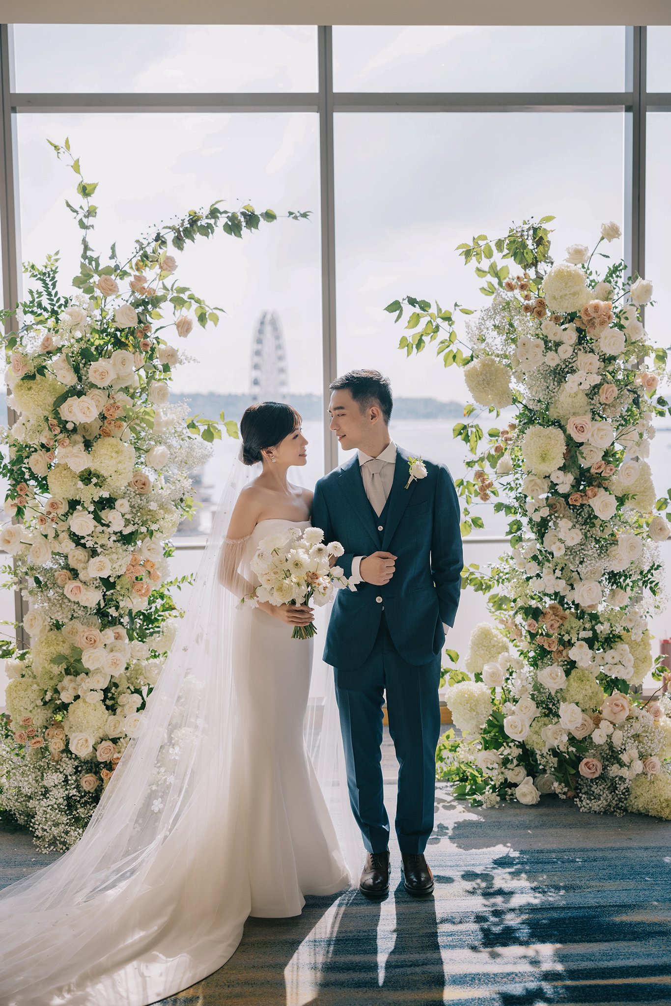 Bride and groom standing in front of a large window with a city skyline in the background, decorated with floral arrangements.