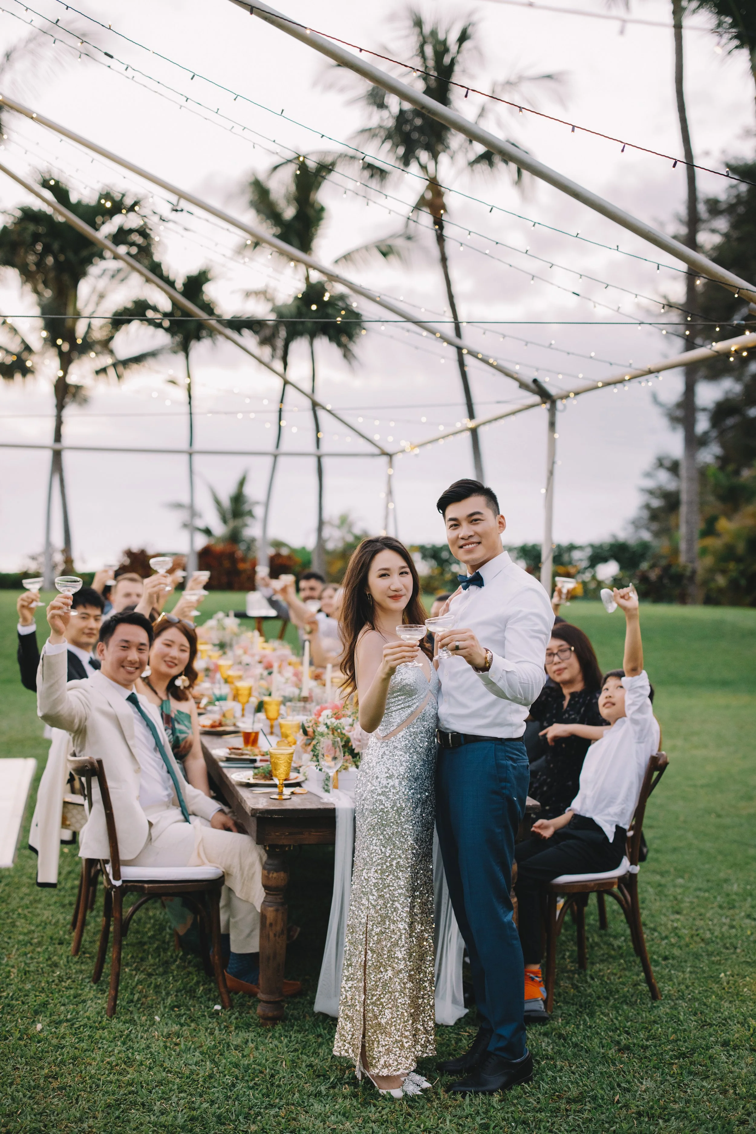 A group of people celebrating at a dinner party outdoors with string lights, palm trees, and a grassy area, as a couple in front toasts with drinks.