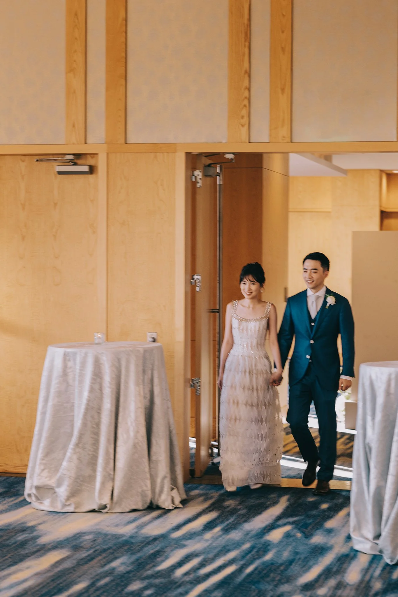 A couple dressed in formal attire, holding hands and smiling, walking out of a wooden doorway in a decorated venue.