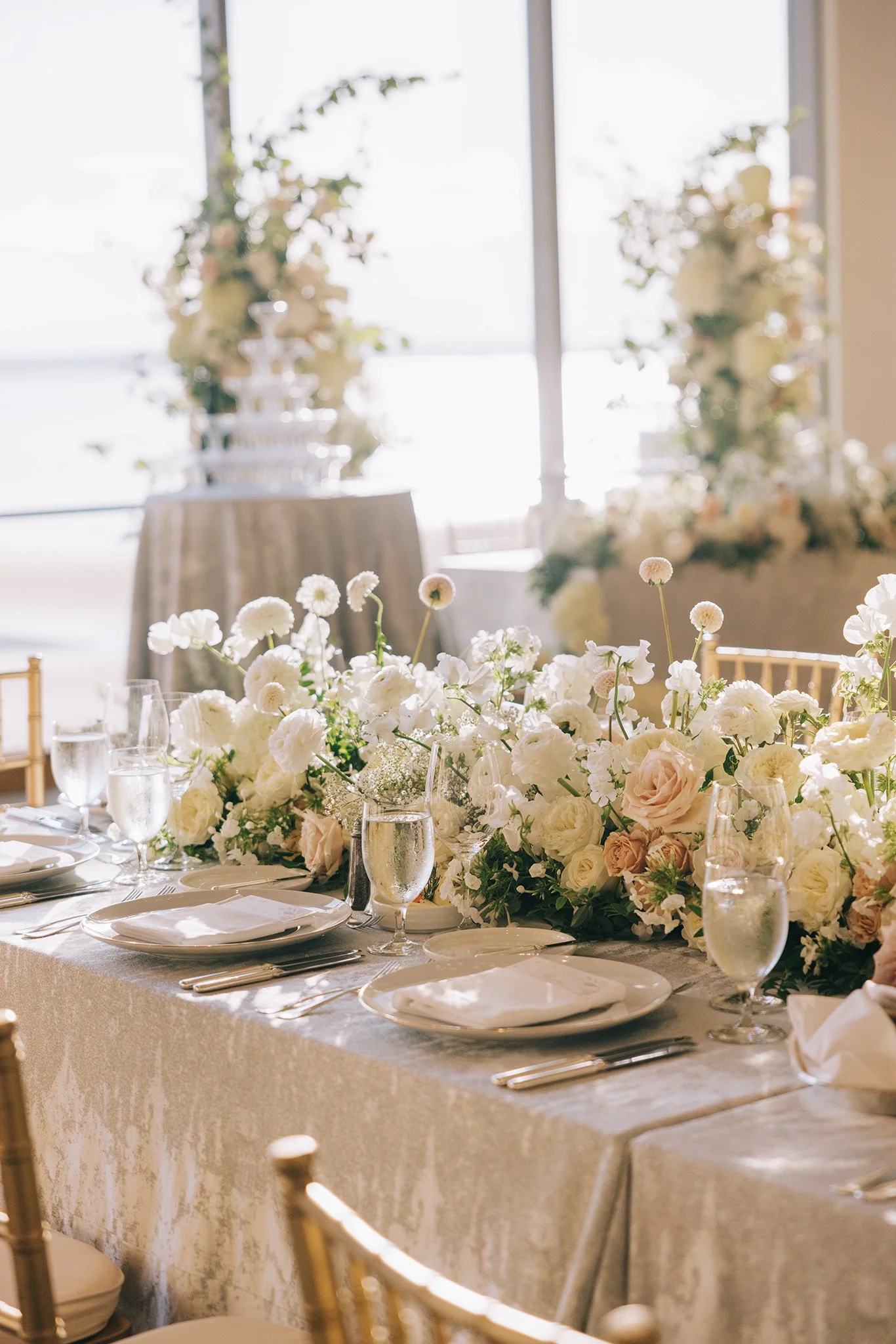 Elegant wedding reception table decorated with white and blush roses, ranunculus, and baby's breath, set with gold and white tableware, leading to a floral and champagne display near a window with natural light.