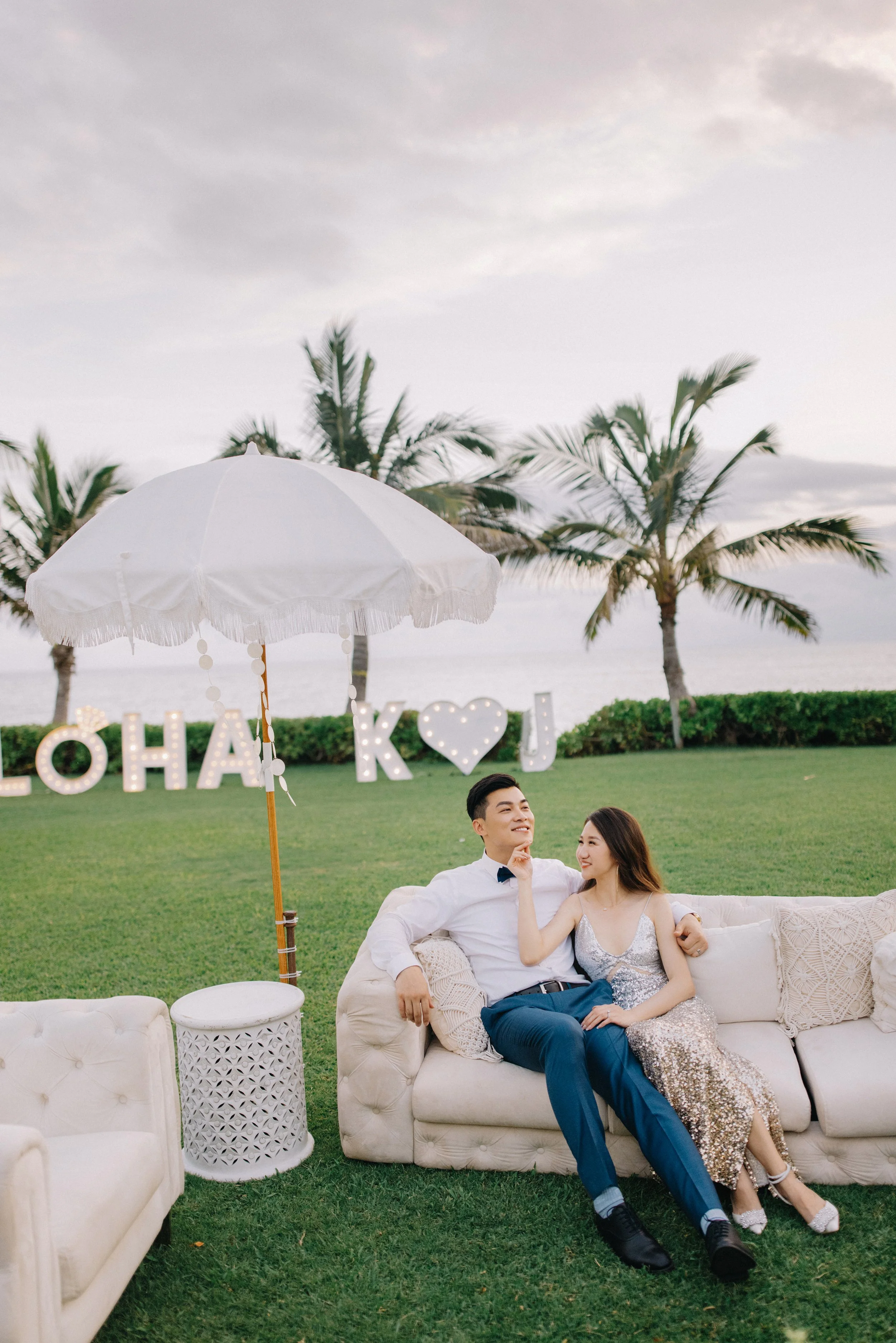 A smiling couple is sitting on a cream-colored sofa outdoors on a grassy area near palm trees and the ocean, with large illuminated letters spelling 'ALOHA' and 'J' and a heart symbol in the background, under a cloudy sky.