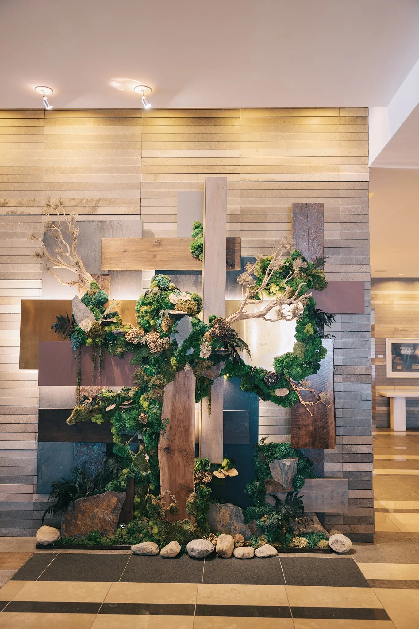 A decorative wall installation featuring a large wooden cross entwined with green moss, plants, and branches, set against a textured brick wall in an indoor space.