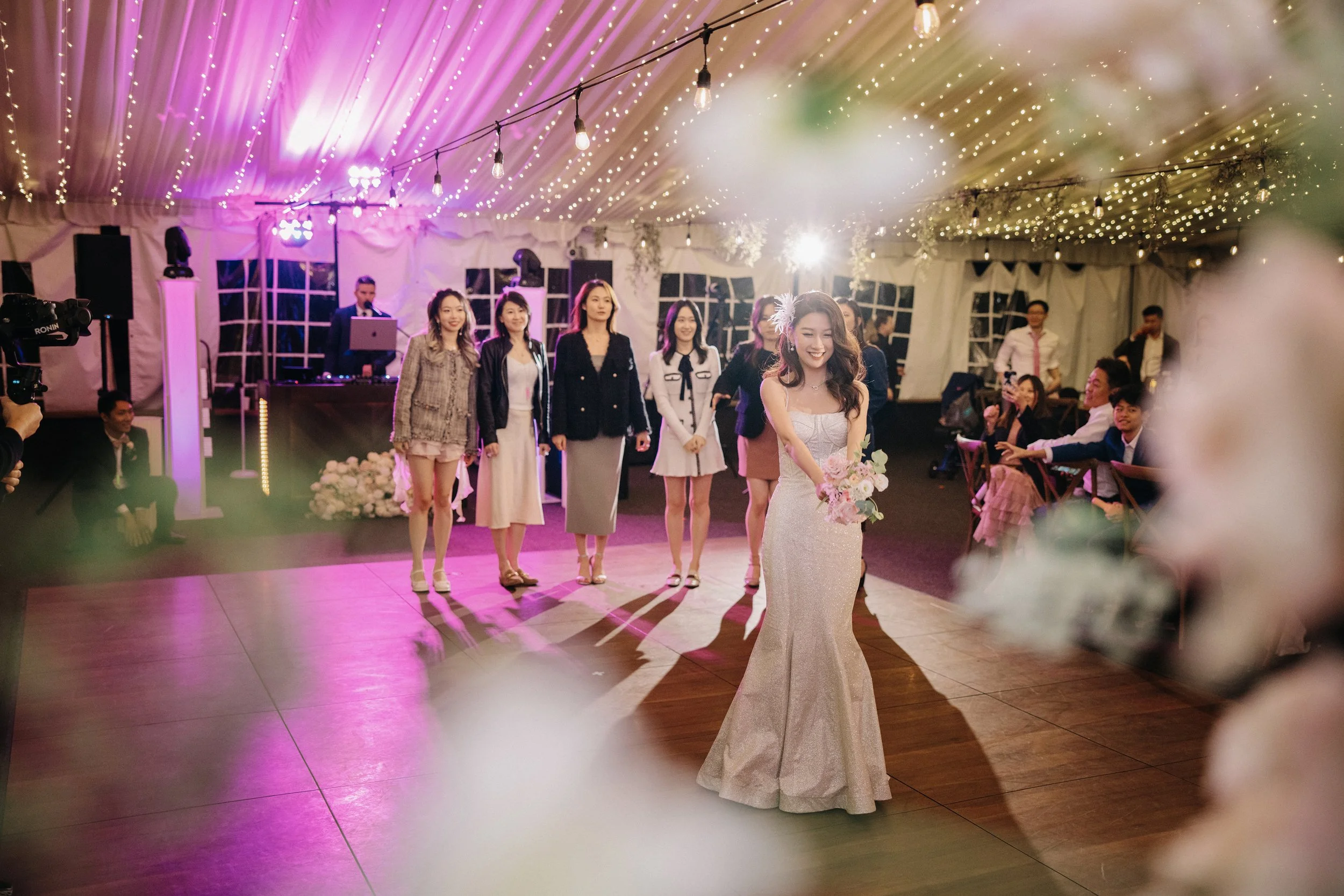 A bride in a silver gown holding a bouquet stands on the dance floor during her wedding reception, with friends and guests behind her and a DJ in the background under a decorated tent with string lights.