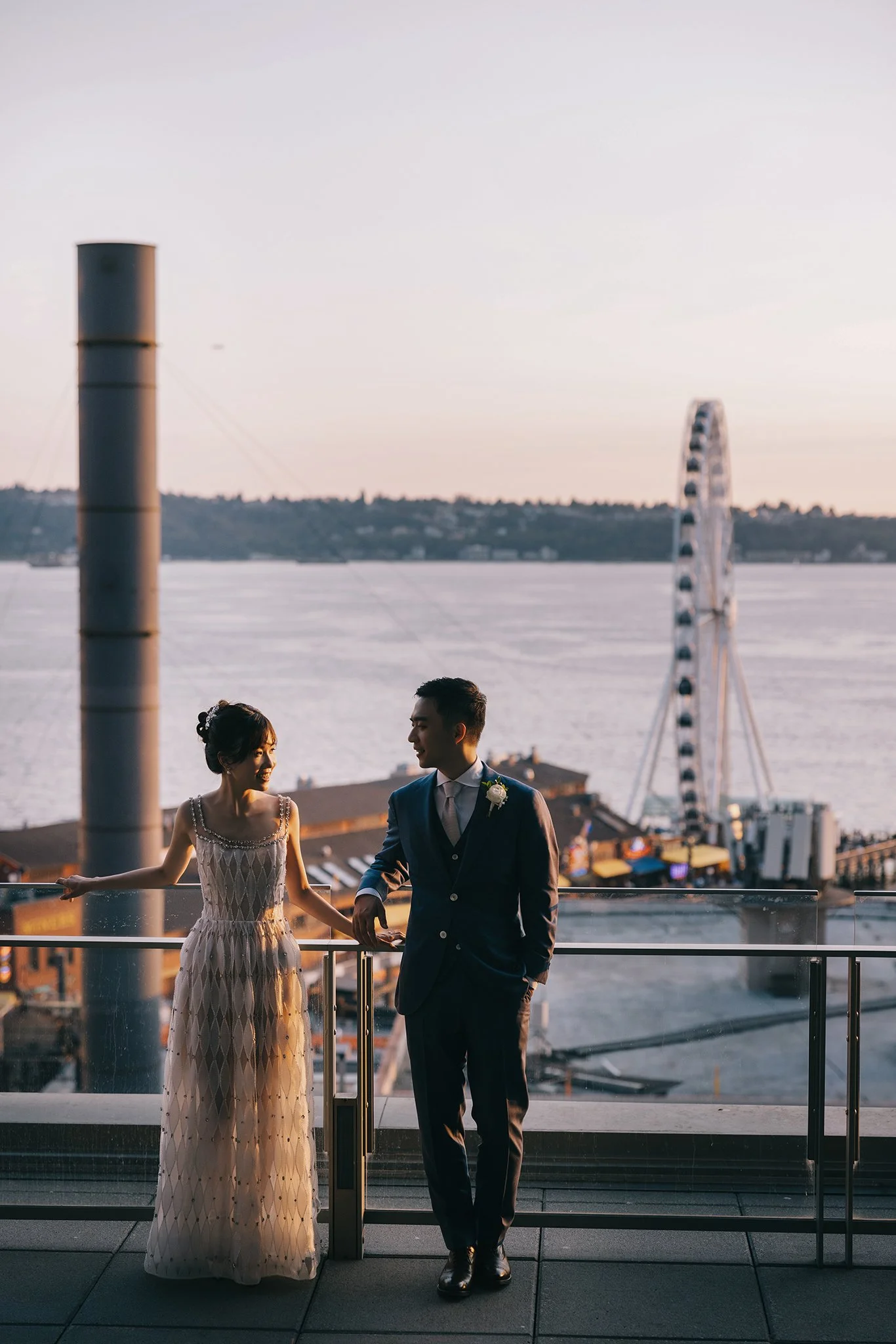 A couple dressed in wedding attire on a balcony overlooking a body of water, with a ferris wheel in the background during sunset.