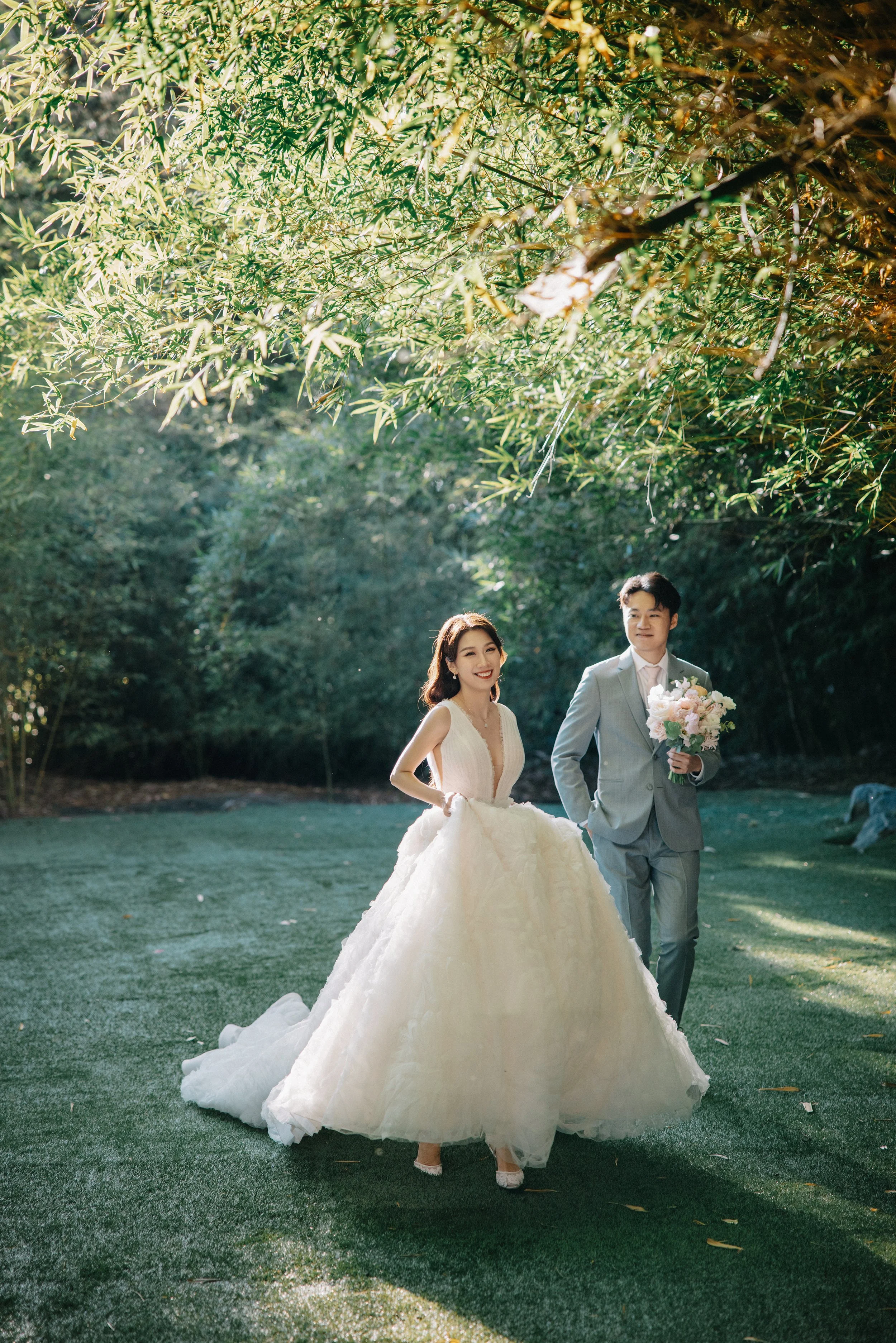 A bride in a white wedding gown and a groom in a light gray suit walk outdoors under green trees, with the bride smiling and the groom holding a bouquet of flowers.