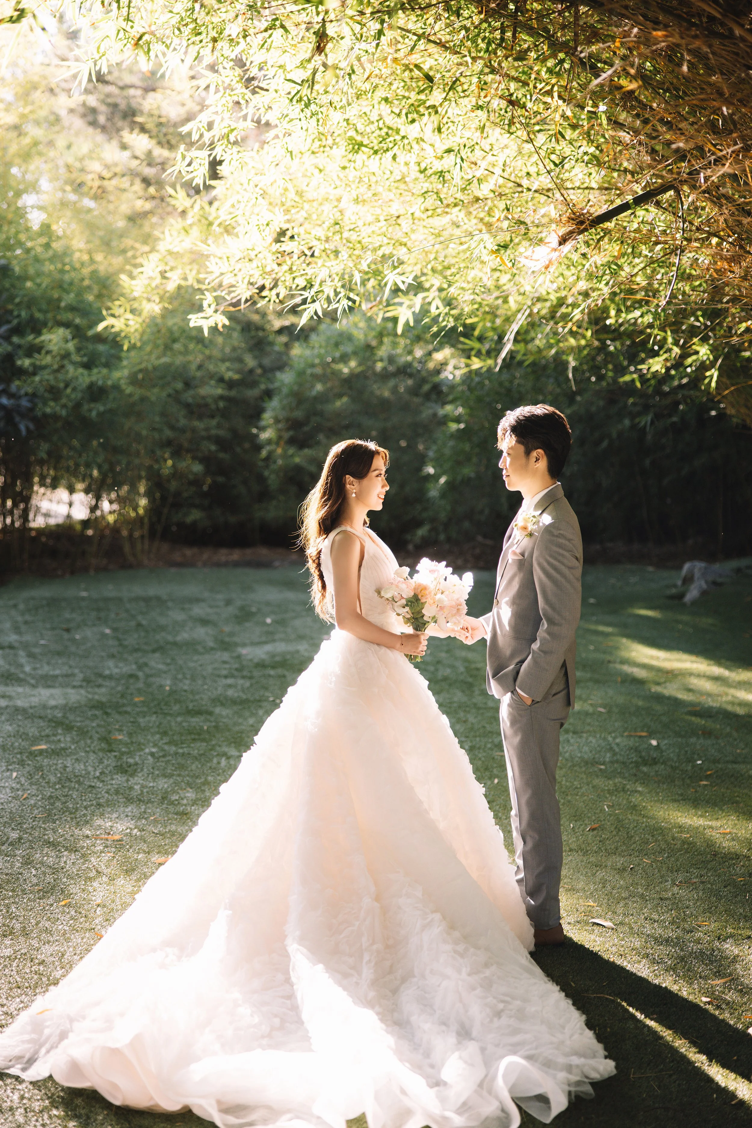 A bride and groom standing outdoors in sunlight, holding hands, with the bride holding a bouquet of flowers. The bride is wearing a white wedding gown with a long train, and the groom is in a light gray suit with his hand in his pocket.