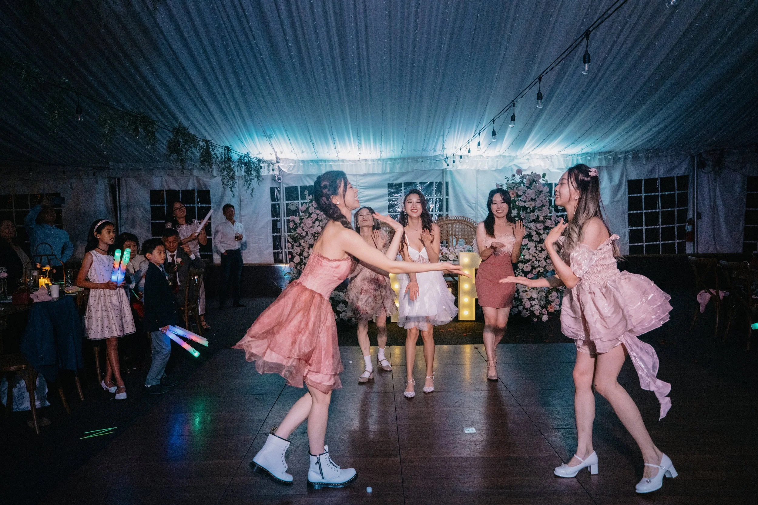 Women dancing at a celebration in a decorated tent with floral arrangements and string lights.