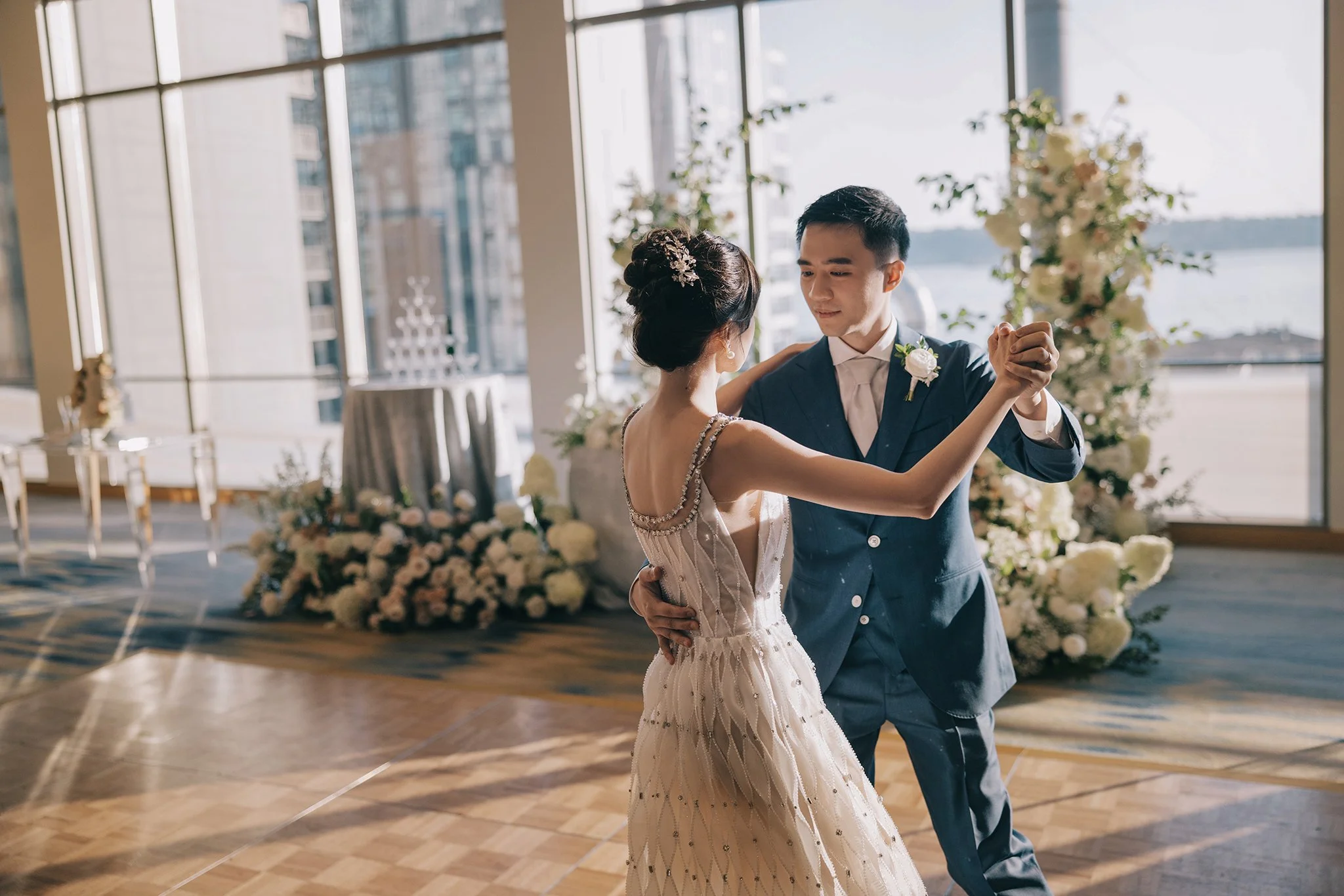 An Asian bride and groom dancing at their wedding reception with large windows and floral decorations in the background.