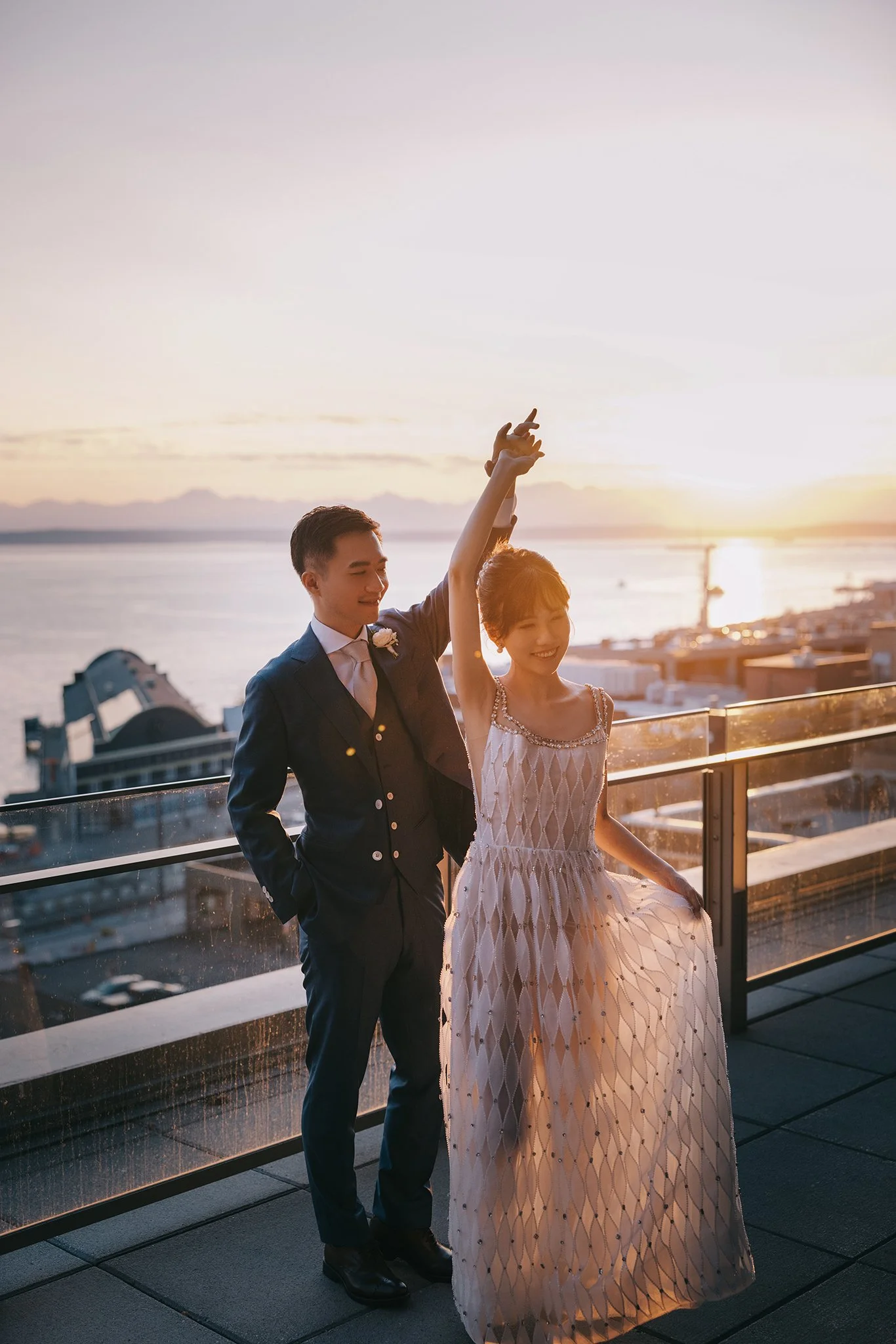 A couple dancing on a rooftop balcony during sunset, with the ocean in the background. The woman is wearing a white dress and the man is in a dark suit.