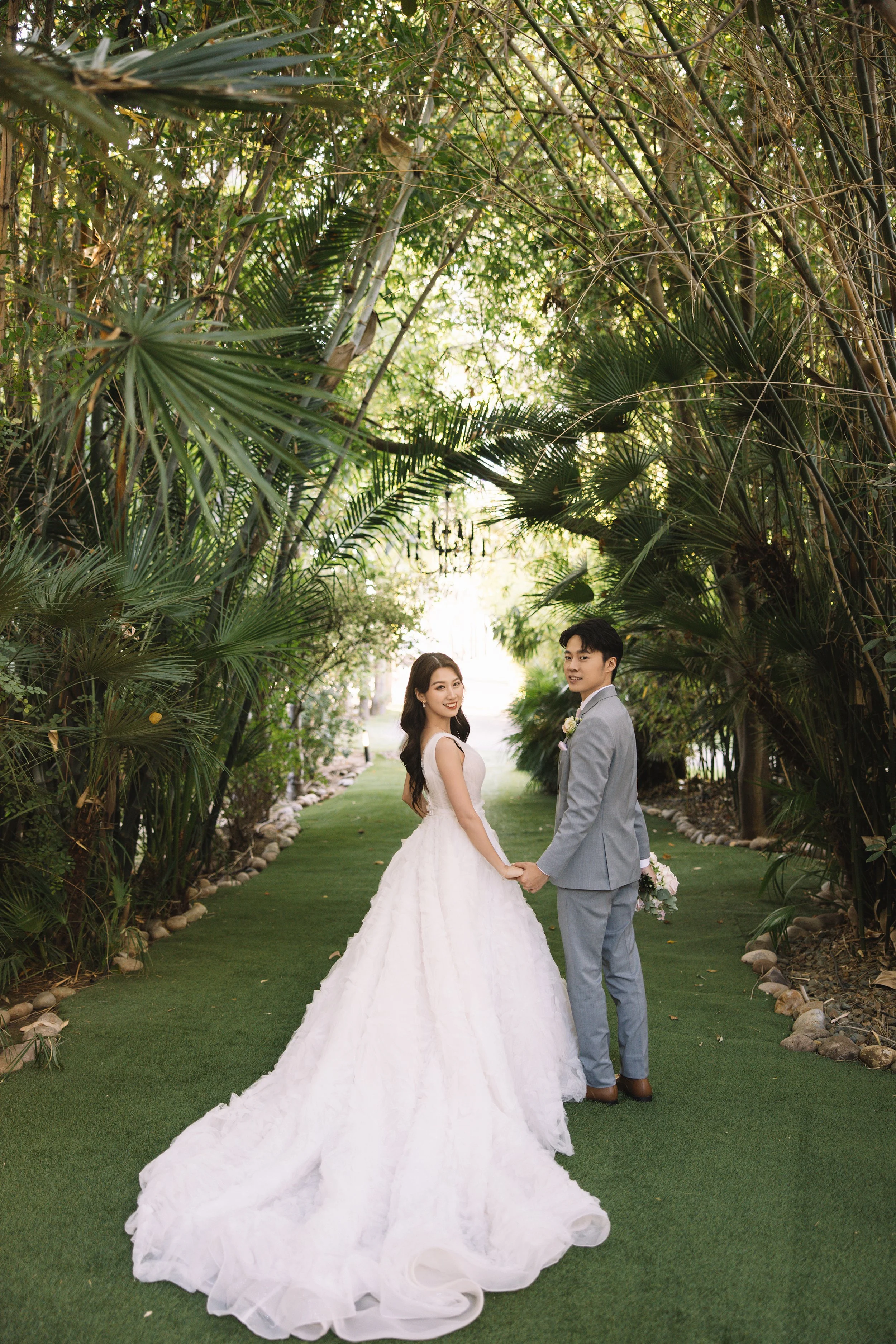 Bride and groom holding hands in a lush green garden archway, with the bride in a white wedding gown and the groom in a light gray suit, park pathway in the background.