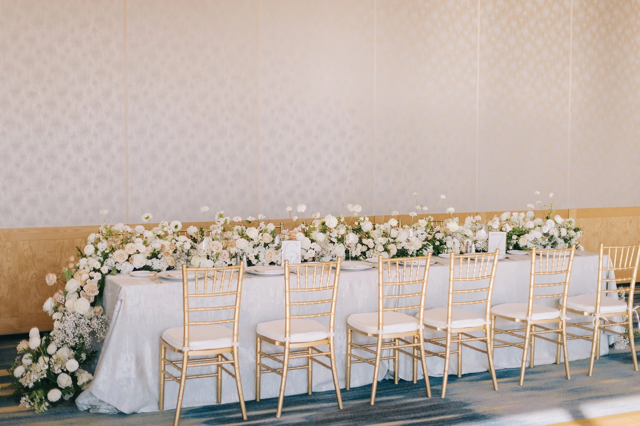 Long banquet table decorated with white roses, baby's breath, and greenery, set for an event, with gold chairs and tableware, in an elegant indoor setting.