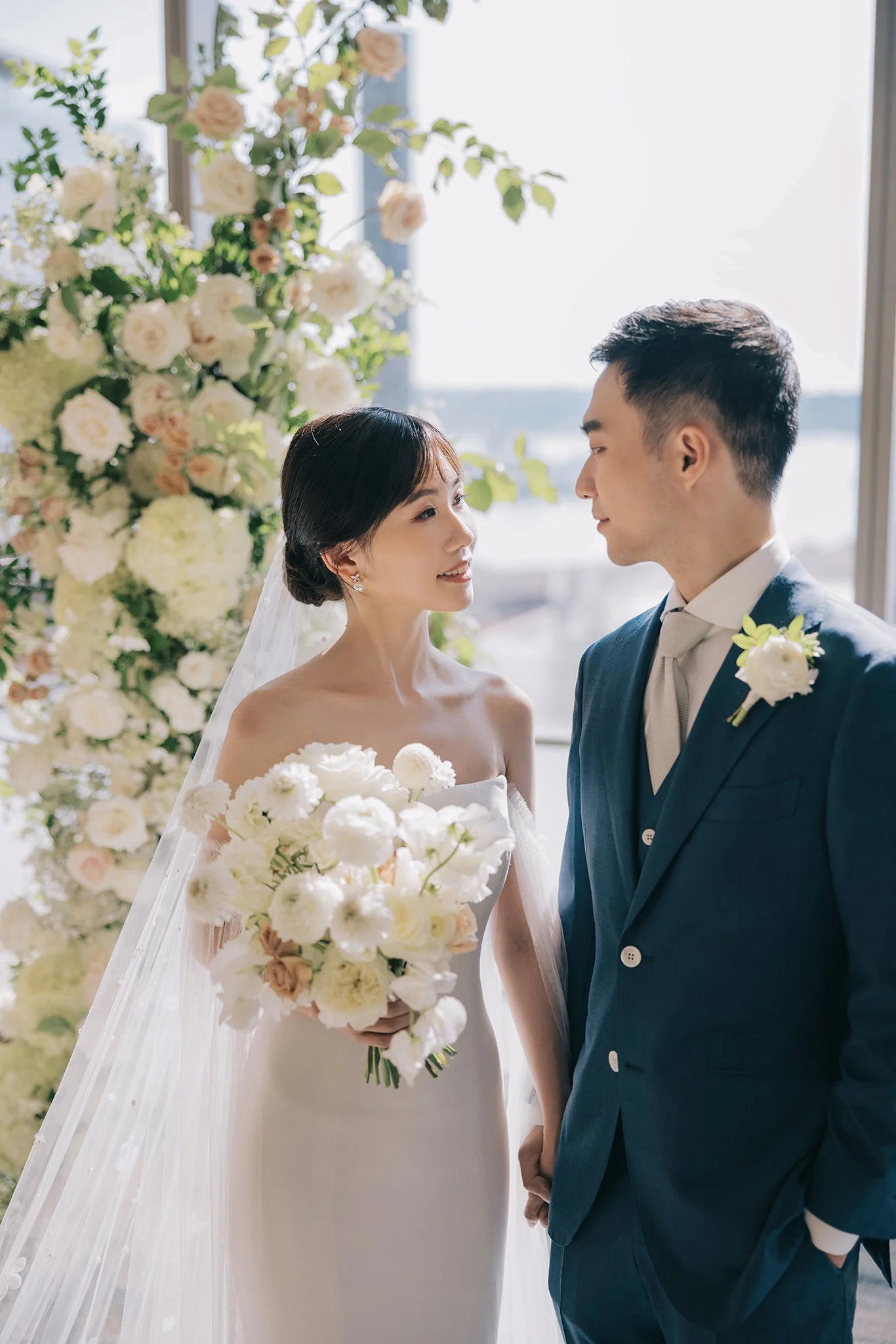 A bride and groom standing indoors during their wedding ceremony, facing each other near a large window with a floral arrangement in the background. The bride is holding a bouquet of white flowers, wearing a strapless white wedding gown and veil. The