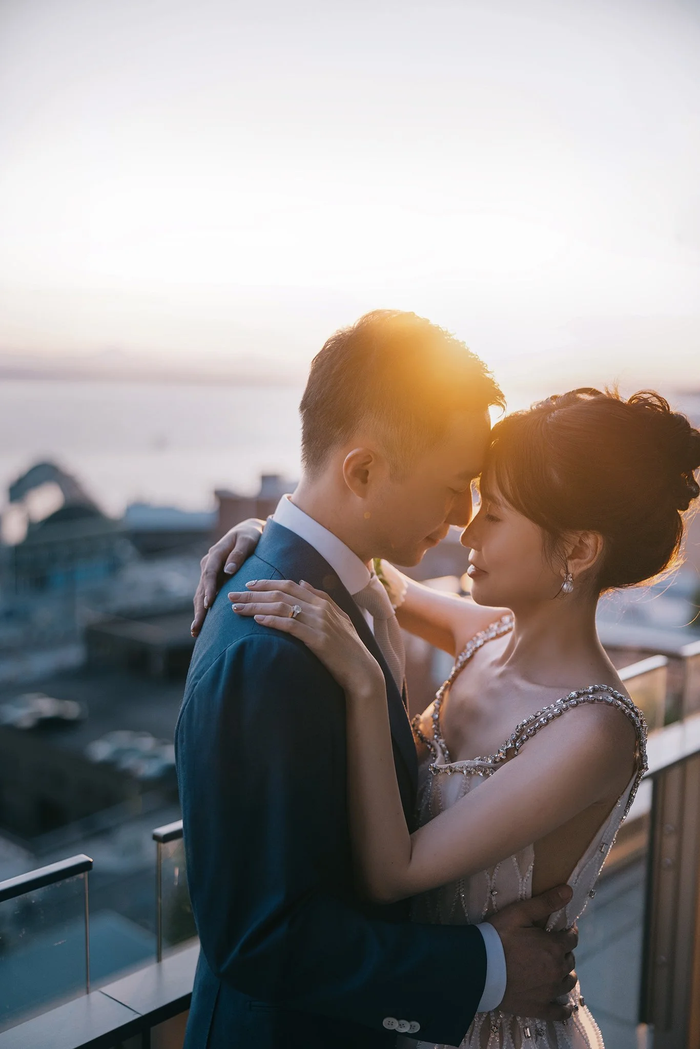 A couple dressed in wedding attire embrace on a balcony with a sunset and cityscape in the background.