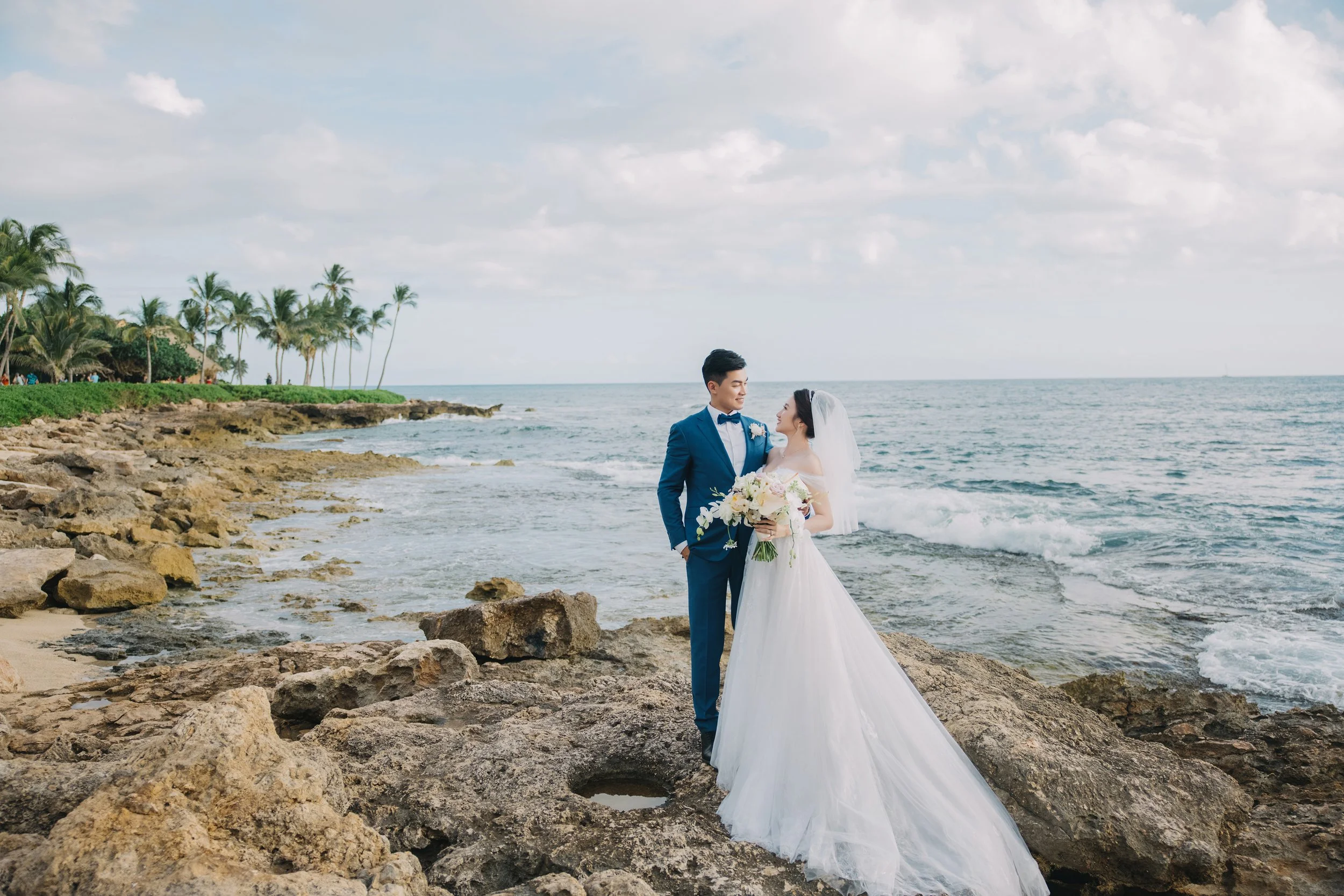 A bride and groom stand on rocks by the ocean, dressed in wedding attire, with the bride holding a bouquet of flowers, palm trees in the background, and a cloudy sky overhead.
