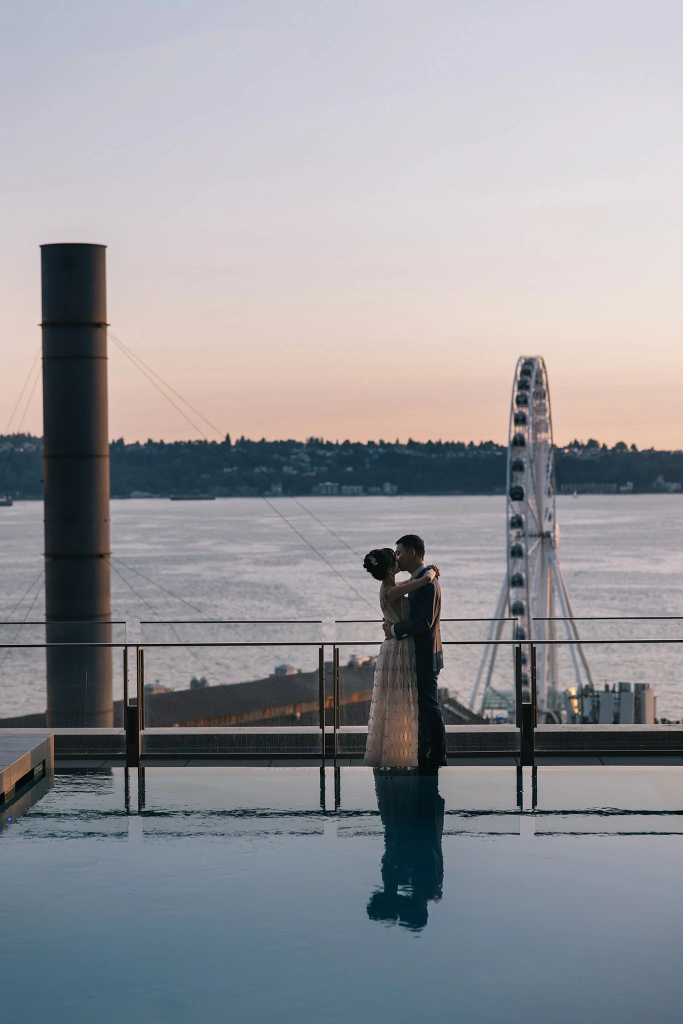 A couple in wedding attire embracing and kissing at sunset by a waterfront pool, with a ferris wheel and body of water in the background.