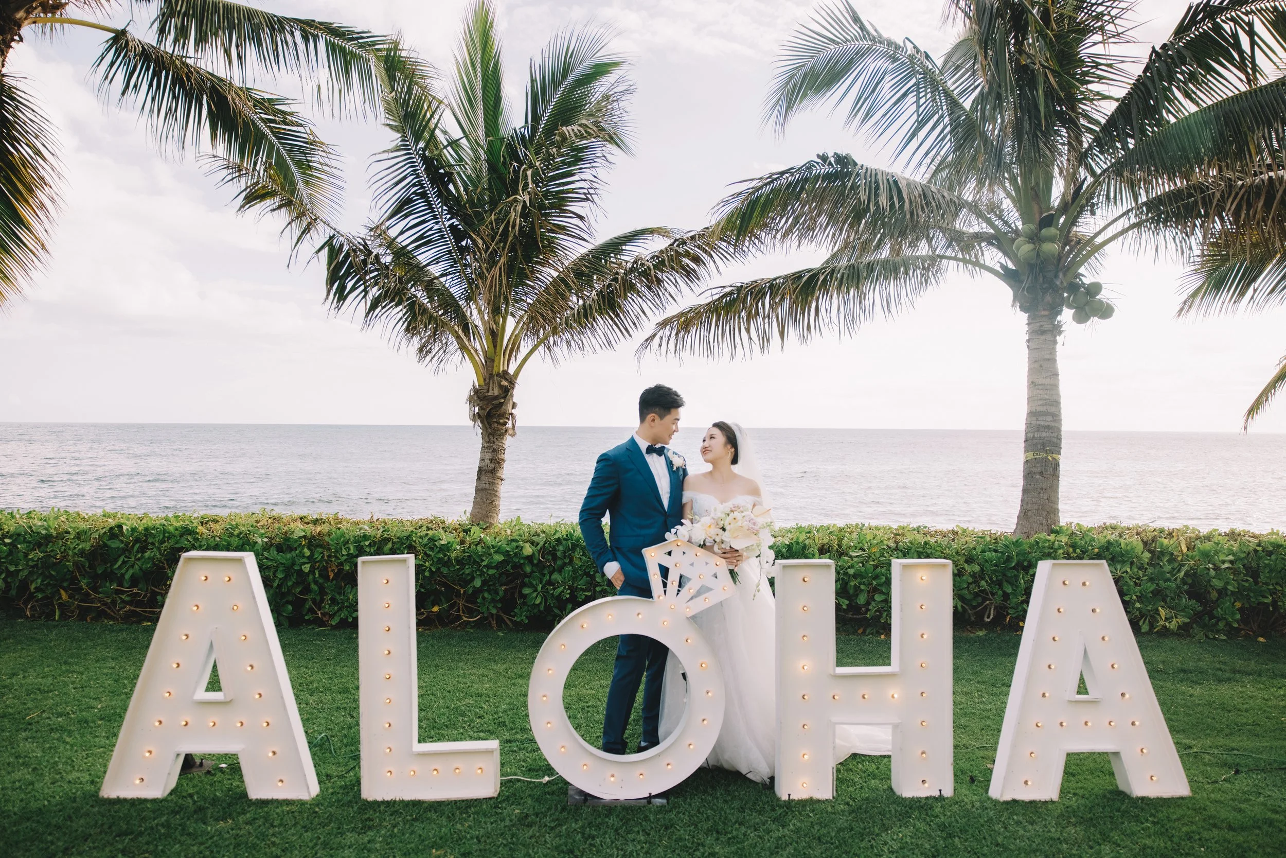 A bride and groom in wedding attire standing outdoors near the ocean, surrounded by palm trees, with large illuminated letters spelling 'ALOHA' in front of them.