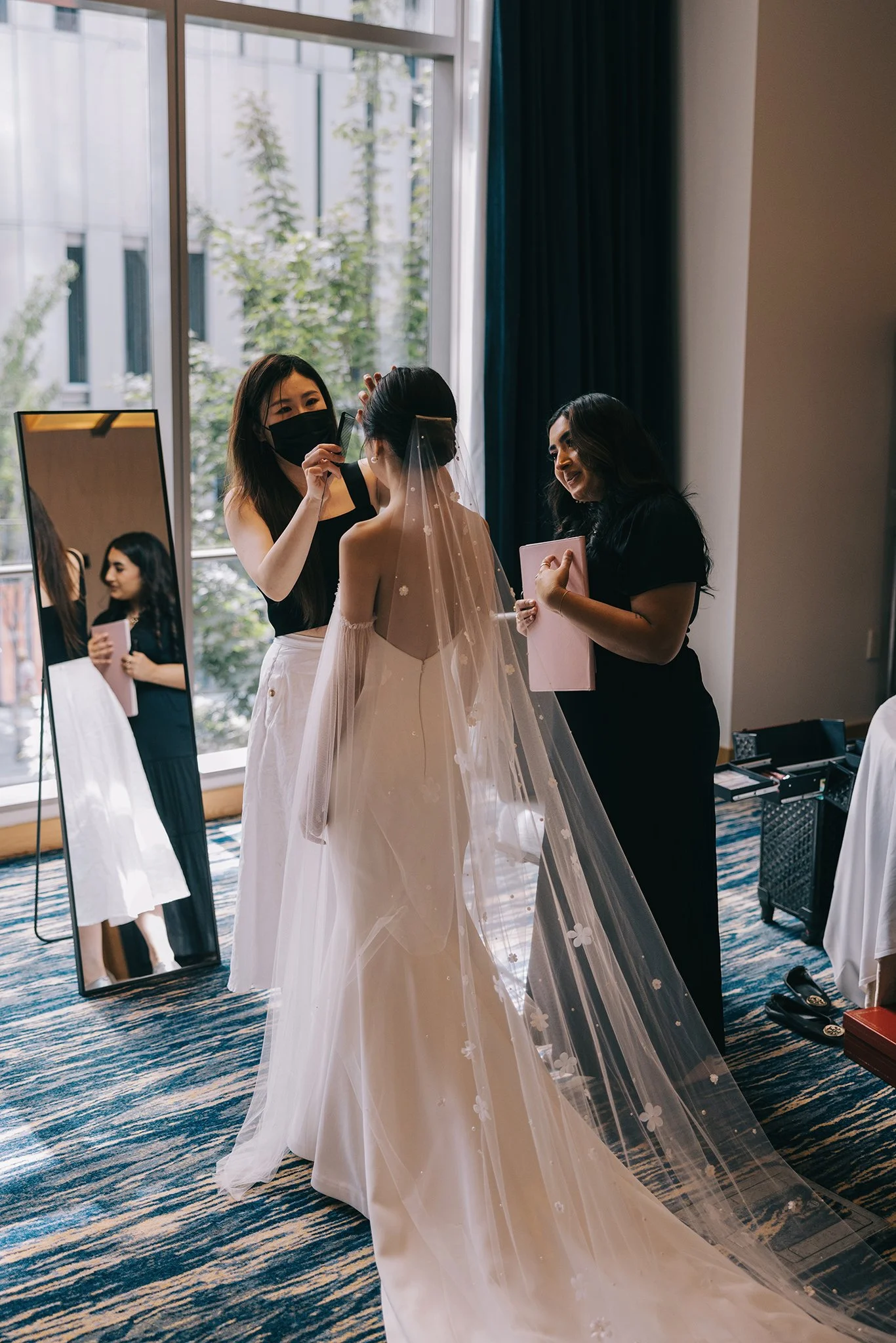 Bridal party having makeup or hair styling session in a hotel room before wedding, with a large window and mirror in the background.