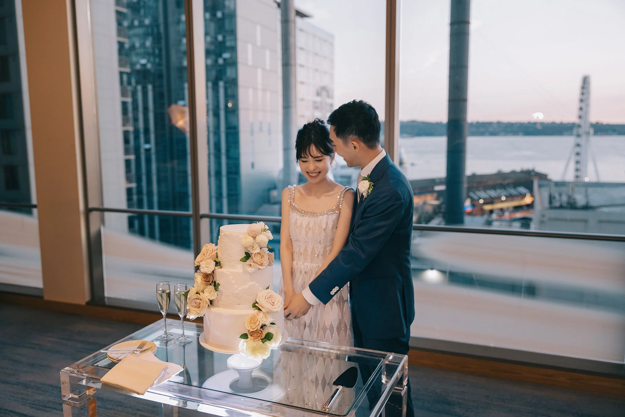 A newlywed couple cutting their wedding cake in a modern venue with large windows overlooking a cityscape and water at dusk.