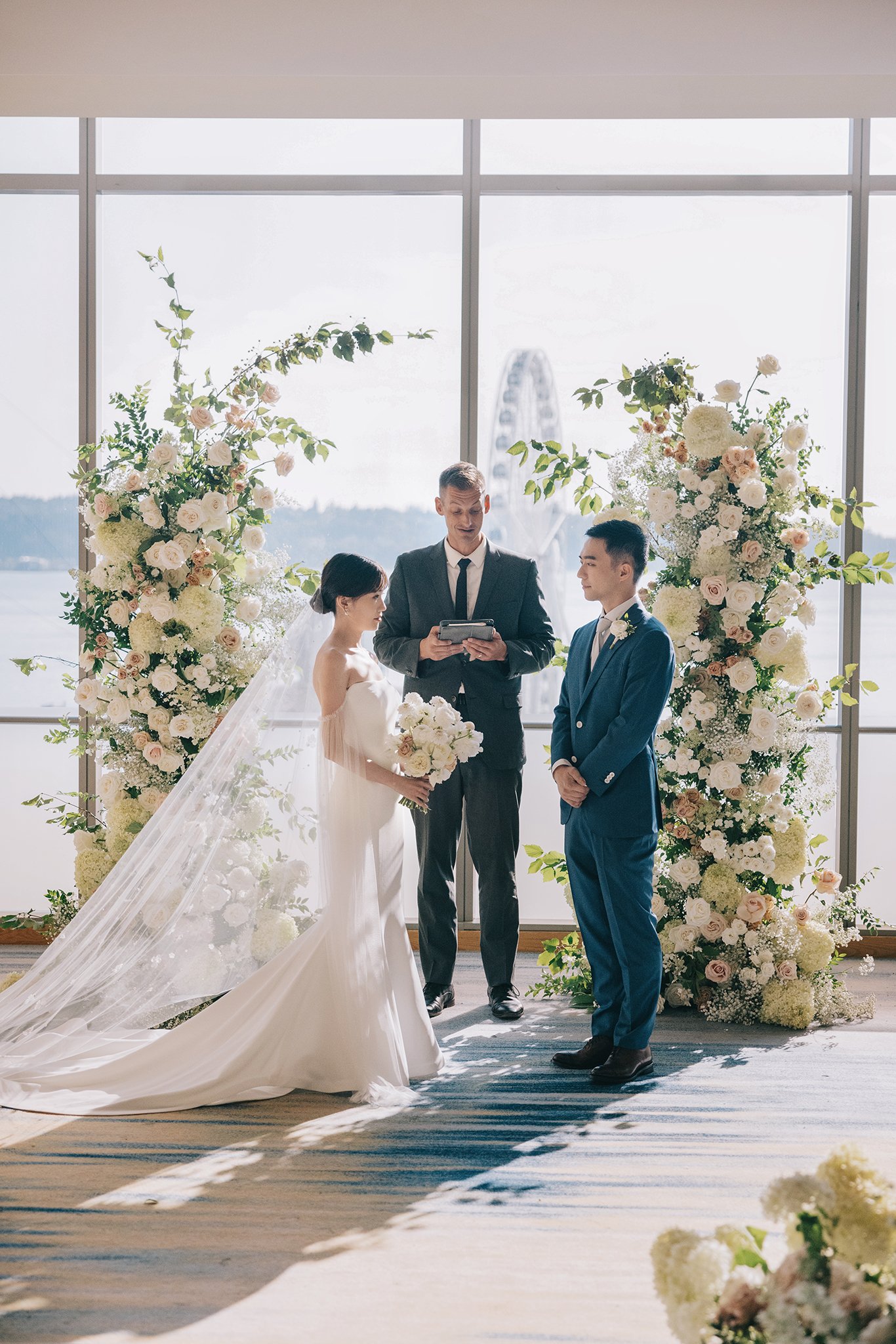 A wedding ceremony with a bride and groom standing before an officiant, surrounded by large floral arrangements, inside a venue with large windows overlooking a waterfront view with a Ferris wheel in the background.
