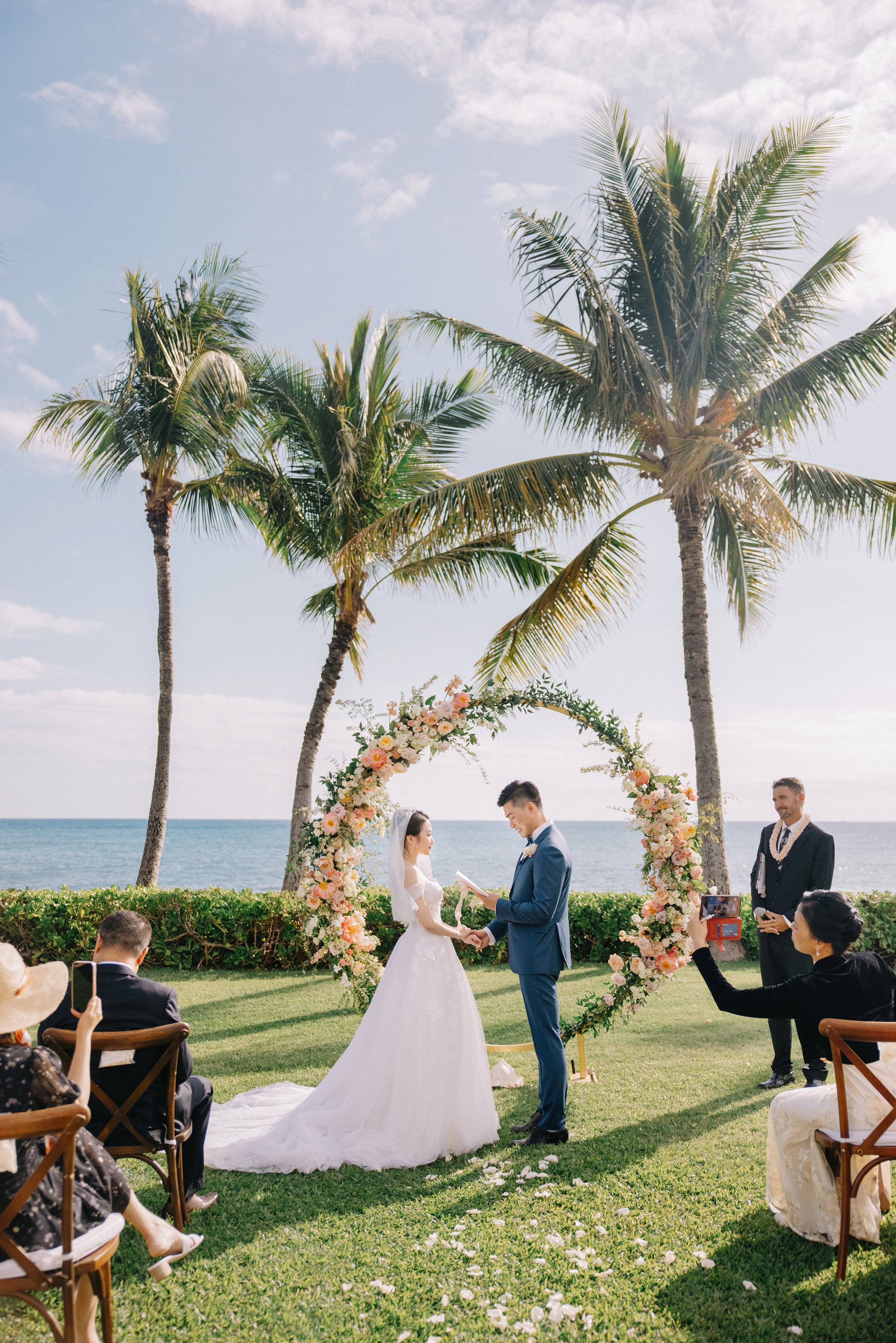 A wedding ceremony taking place outdoors on a grassy area near the ocean, with palm trees and a floral arch in the background. A couple is exchanging vows, with guests seated and one person recording the event on a phone.