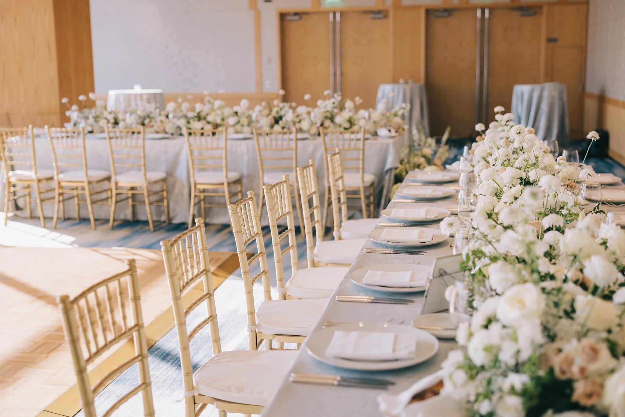 Elegant banquet table setup with white tablecloths, floral centerpieces, and gold chairs in a well-lit event room.