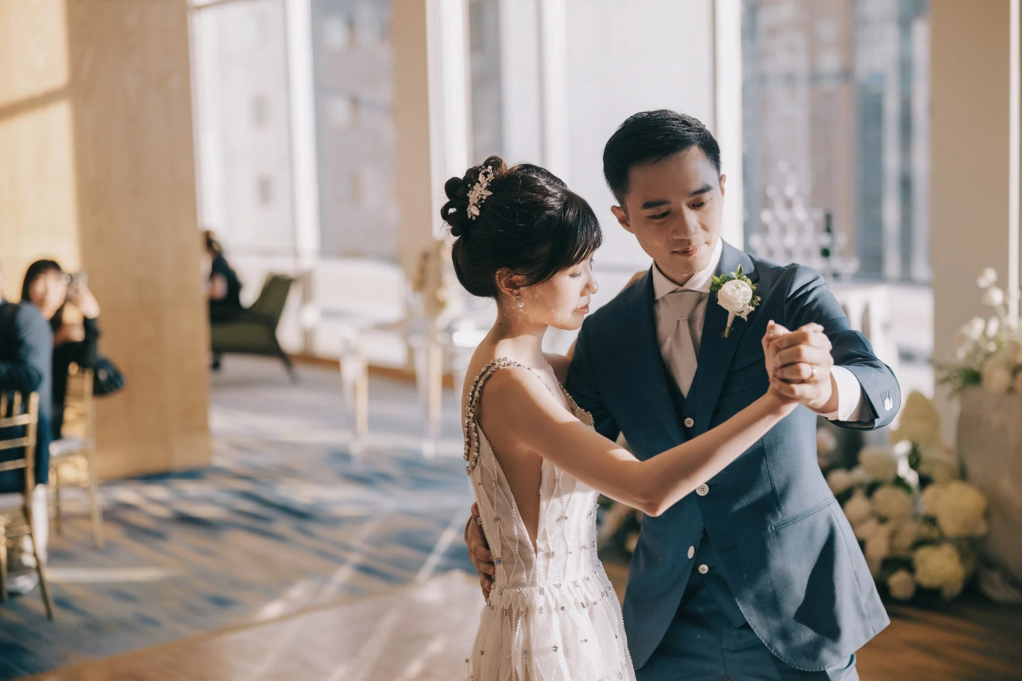 A bride and groom are dancing closely at their wedding reception in a sunlit room with large windows. The bride is wearing an elegant white wedding gown, and the groom is in a blue suit with a white shirt and tie. Other guests are seated and watching