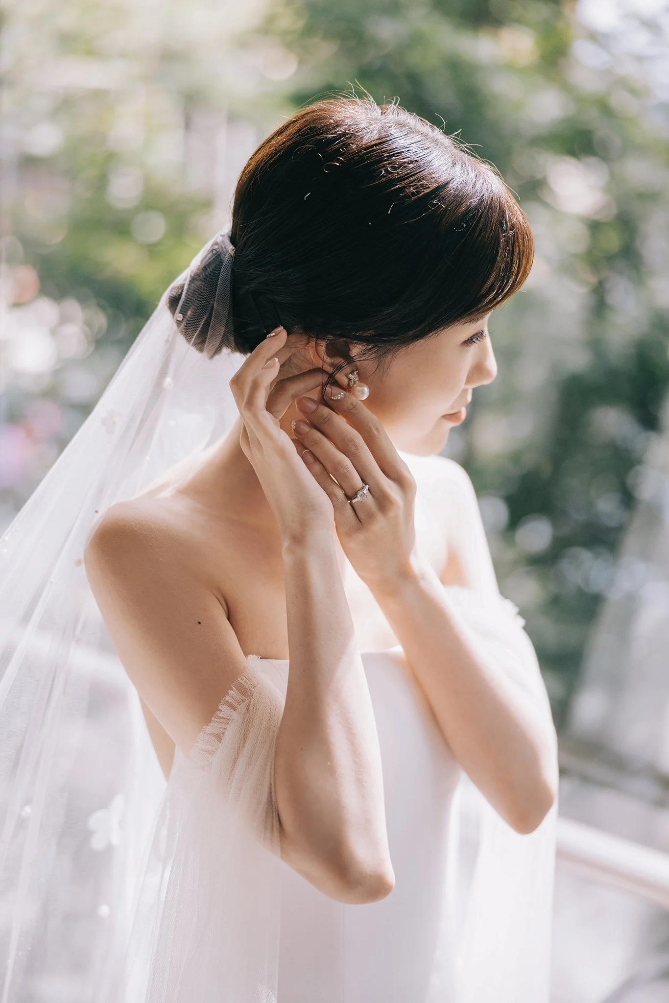 A bride is putting on earrings, wearing a white strapless wedding dress and veil, with a blurred outdoor background.