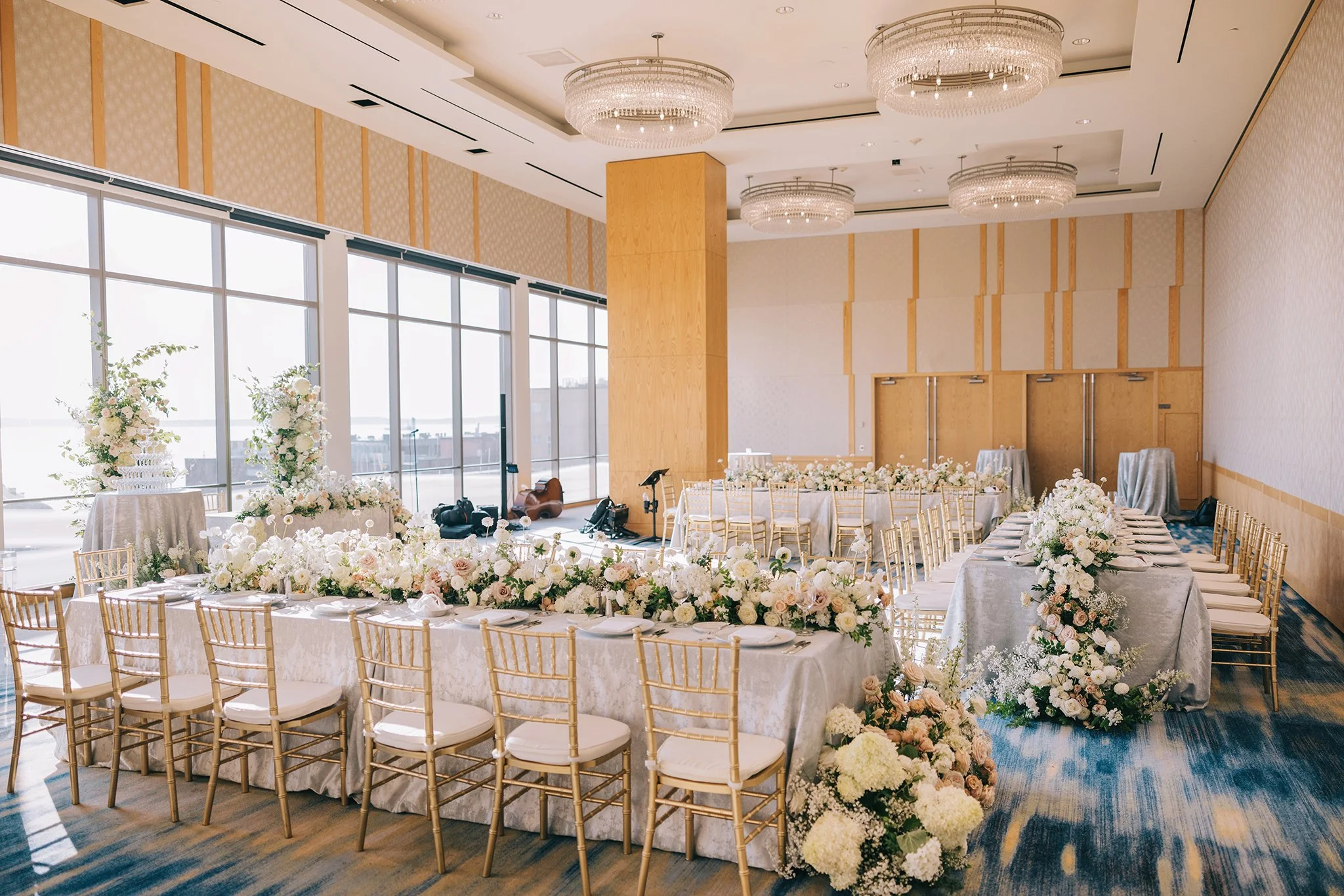 Elegant banquet hall decorated for a special celebration with long tables adorned with white floral centerpieces, gold chairs, and large windows letting in natural light.