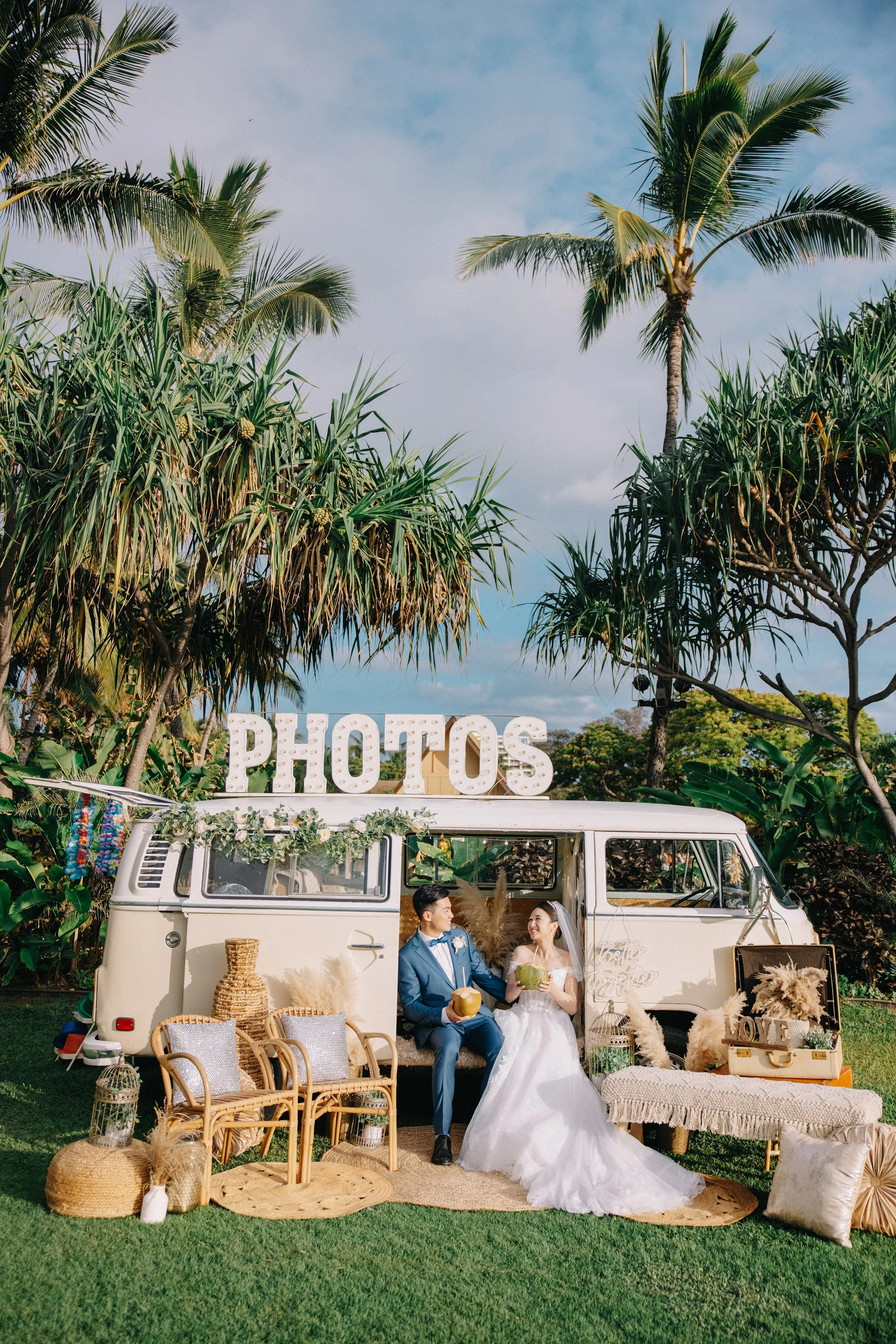 A bride and groom sit in front of a vintage van decorated with flowers, on a lush green lawn surrounded by palm trees. The scene includes wicker chairs, rustic decor, and large letter signs spelling 'PHOTOS'. The bride wears a white wedding gown and 