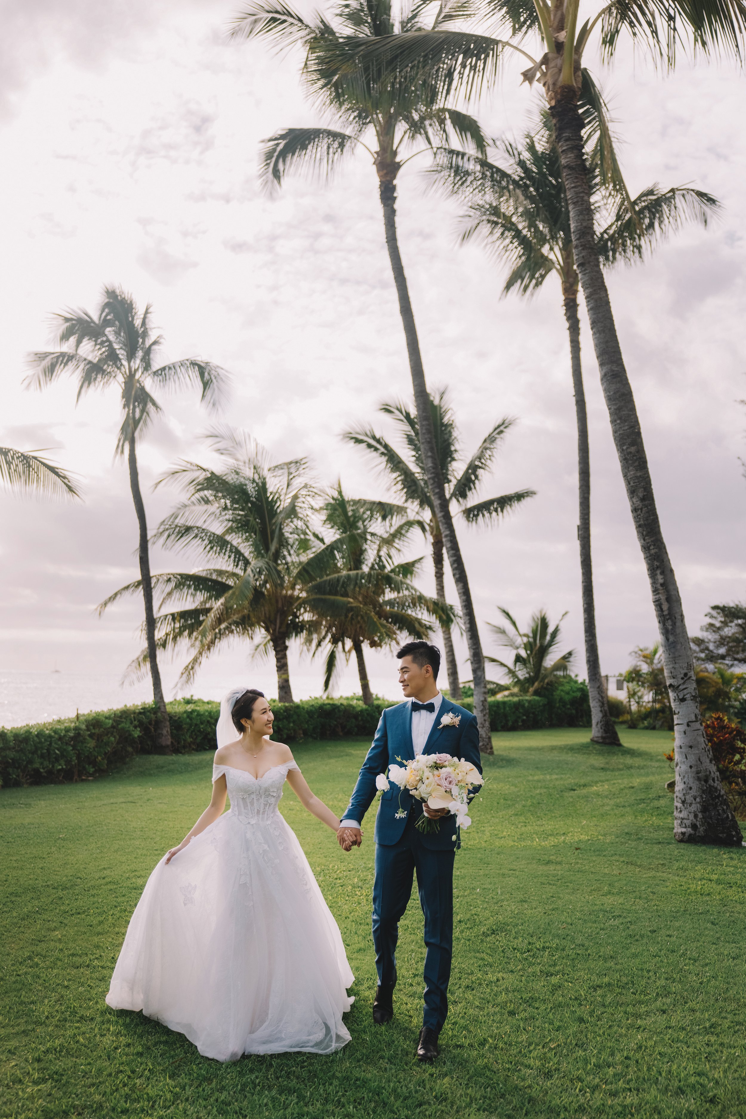 A bride and groom holding hands and walking on a lush green lawn with palm trees and a cloudy sky in the background, at a tropical outdoor wedding.
