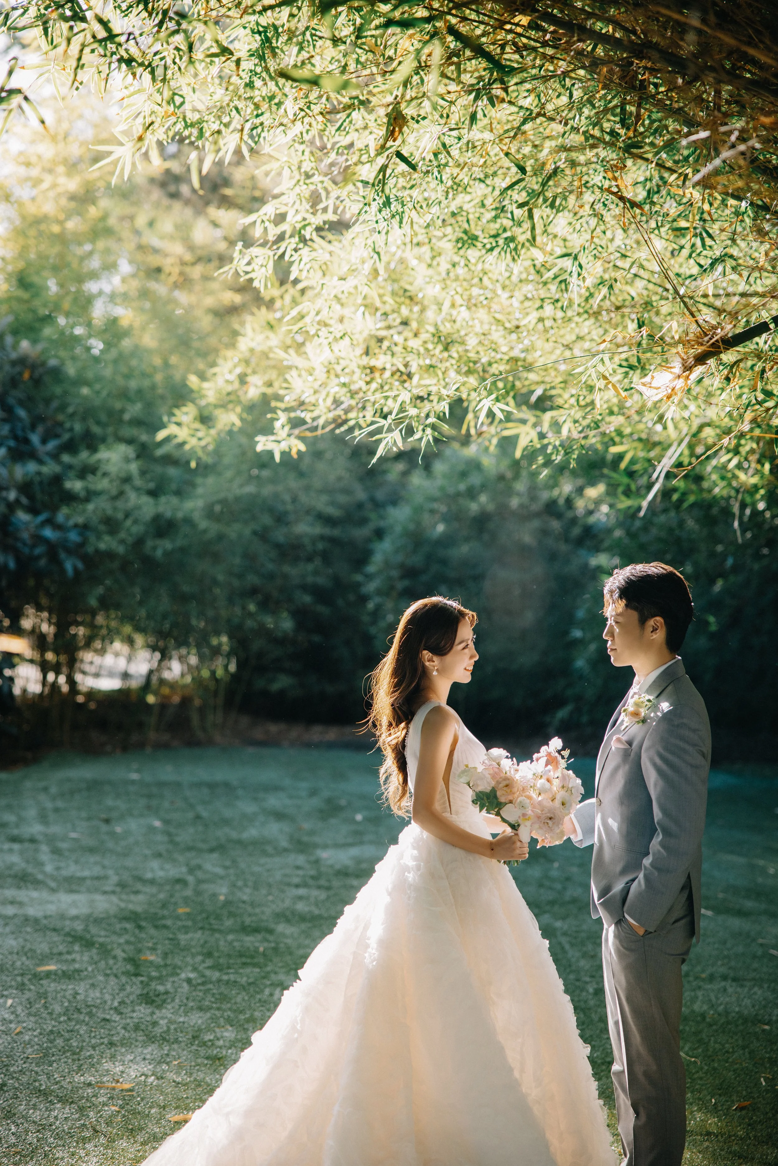 Bride and groom standing face to face outdoors, the bride holding a bouquet of pink and white flowers, surrounded by green trees and sunlight.