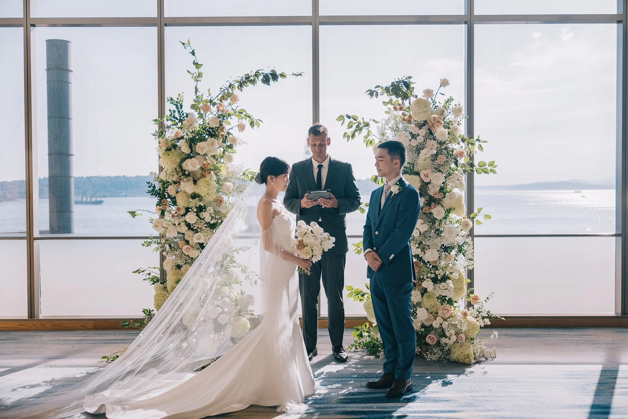 A wedding ceremony taking place indoors with a large window showing water and sky in the background. The bride and groom stand facing each other, with a minister in the middle holding a book. The bride is wearing a white wedding dress with a veil and