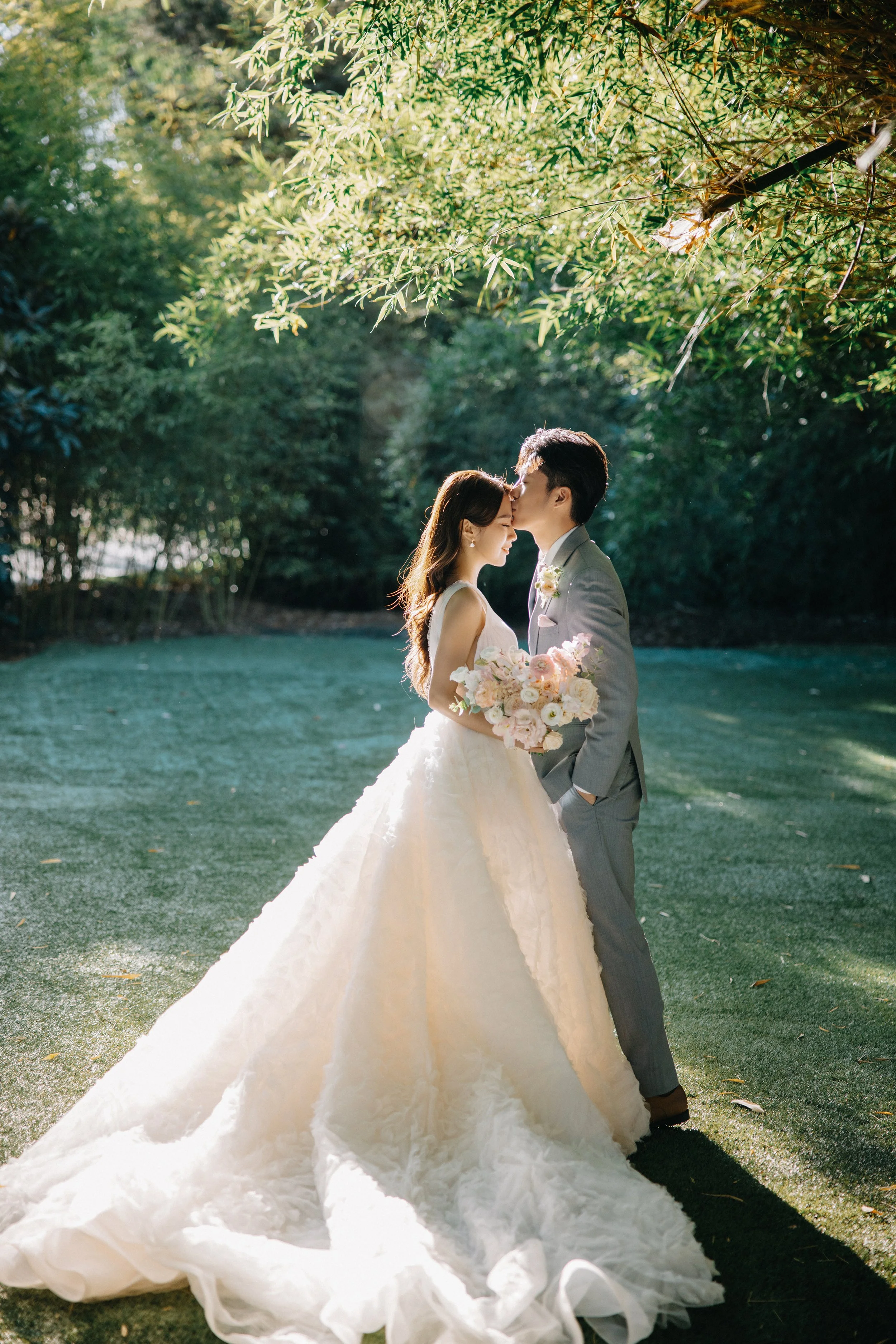 A bride and groom share a kiss in a lush outdoor setting, with greenery overhead and sunlight filtering through the trees; the bride wears a voluminous white wedding gown and holds a bouquet of pale pink and white flowers, while the groom is dressed 