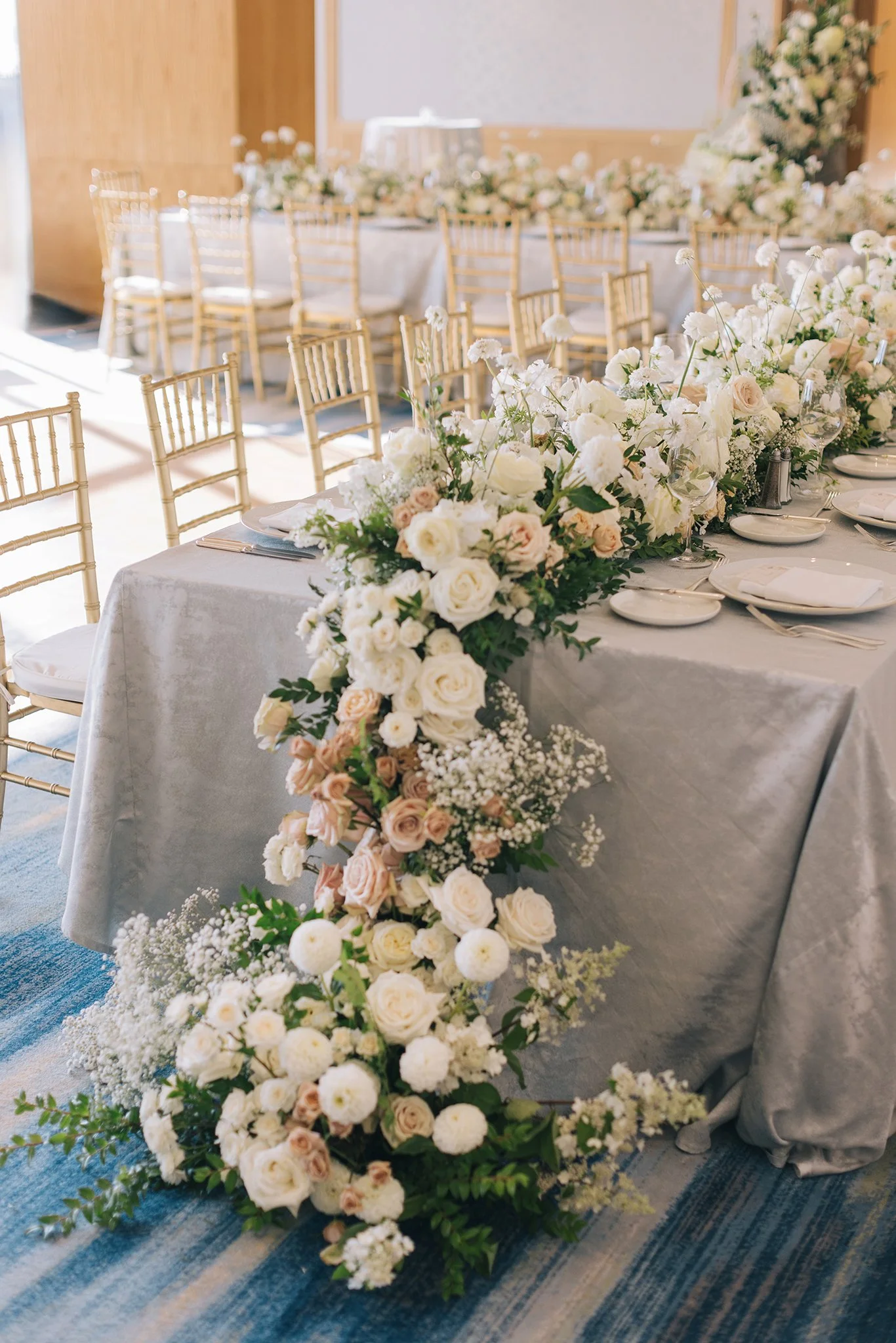Elegant wedding reception table decorated with a layered white and blush floral arrangement, set with white plates, silverware, and napkins, surrounded by gold Chiavari chairs.