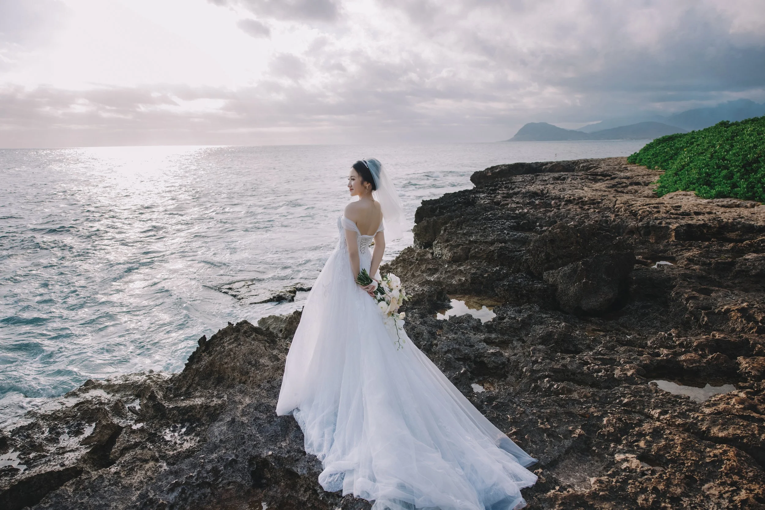Bride in a white wedding gown holding a bouquet, standing on rocky coastline near the ocean with a mountainous island in the distance and cloudy sky overhead.