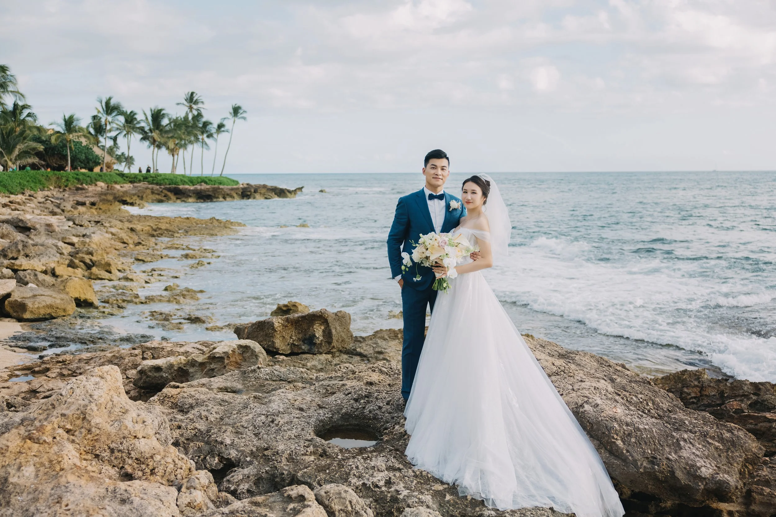Bride and groom standing on rocky shoreline by the ocean, dressed in wedding attire, with palm trees in the background.