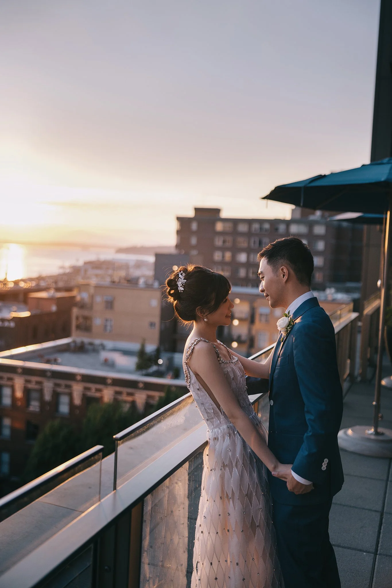 A bride and groom stand on a rooftop balcony at sunset, facing each other and holding hands, with city buildings in the background.